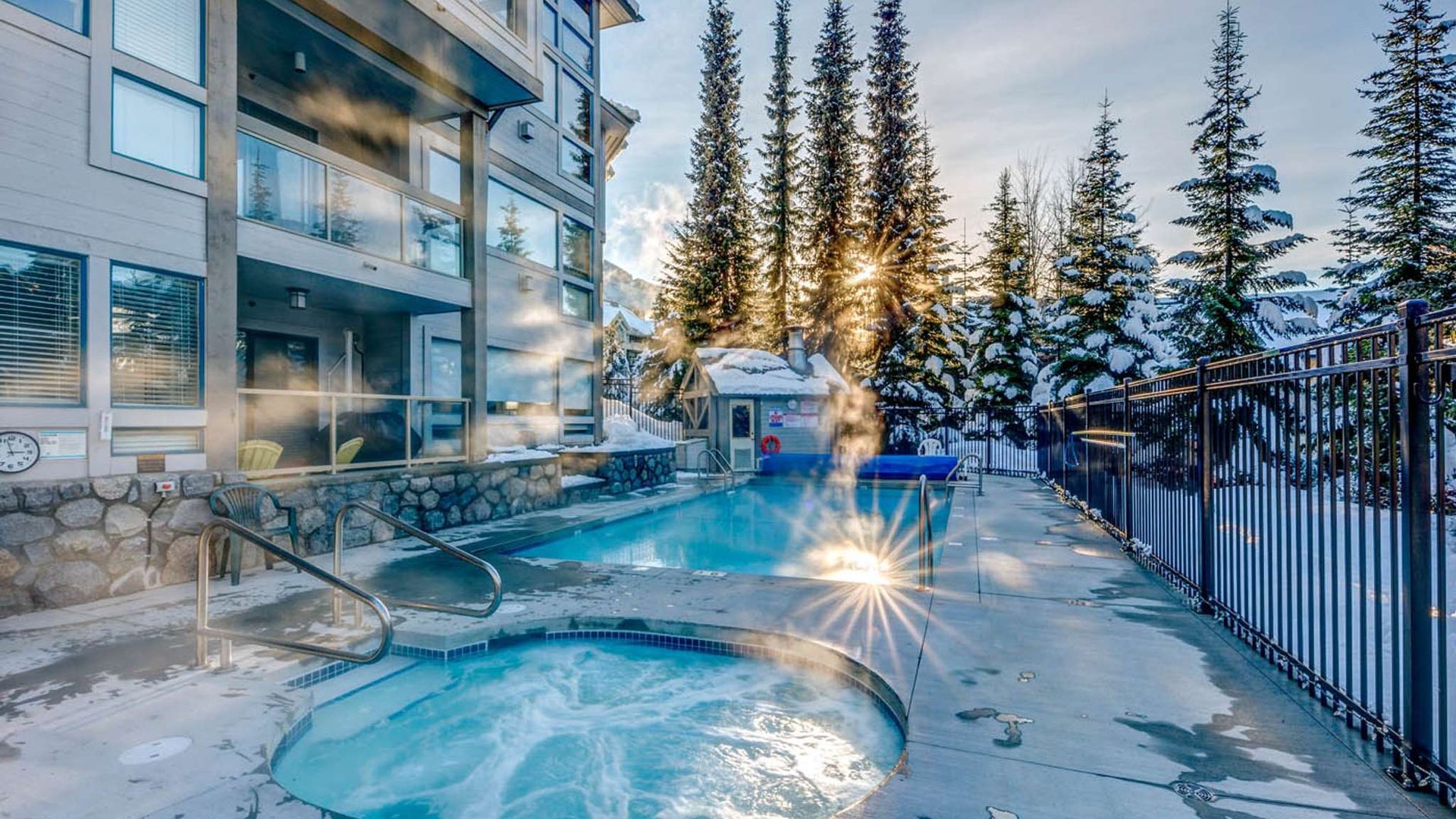 Snowy outdoor hot tub beside a lodge at sunset, with steam, pine trees, and a sparkling light reflection.