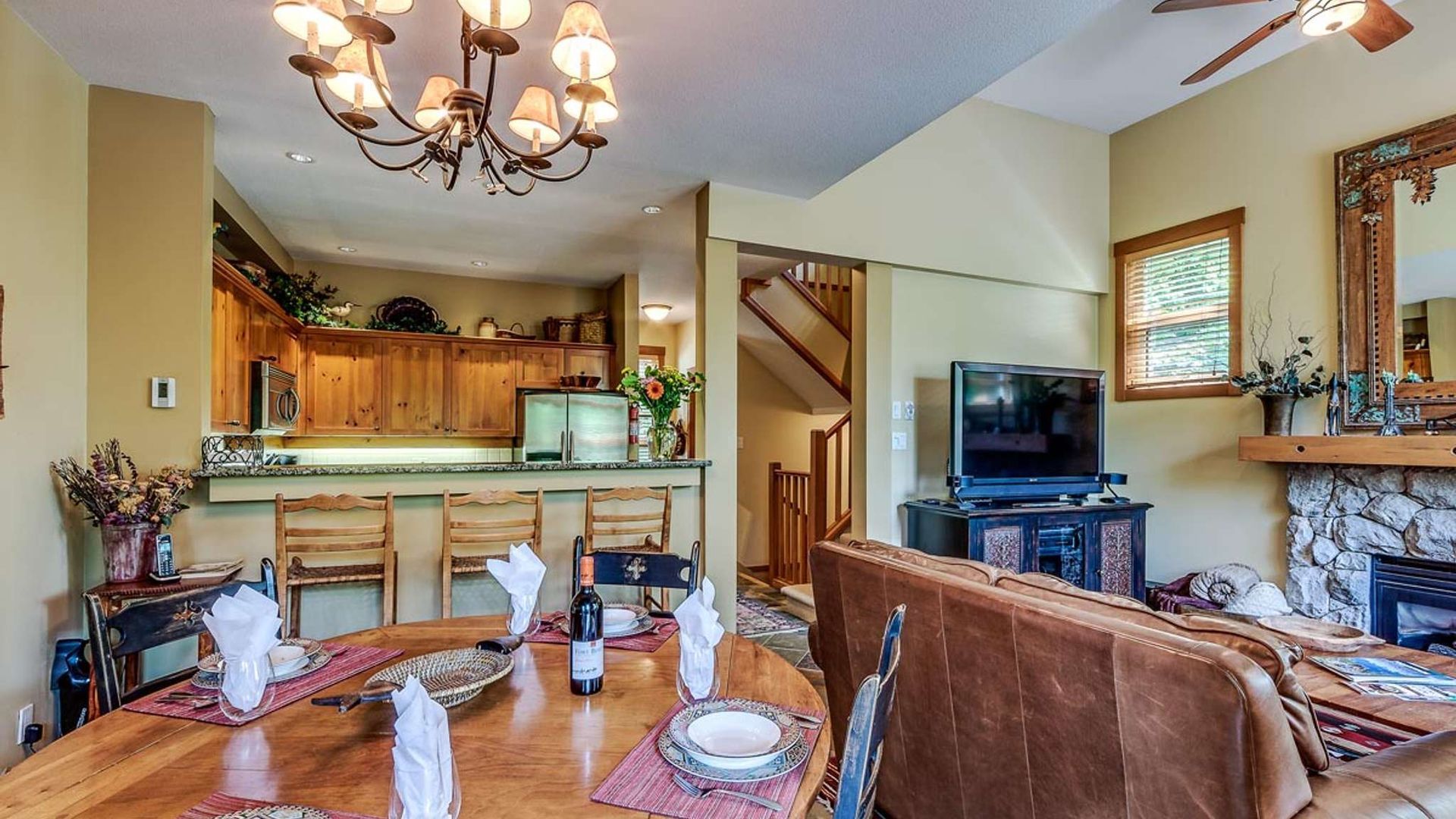 Open kitchen and dining room with wood cabinets, chandelier, stone fireplace, and TV near a staircase