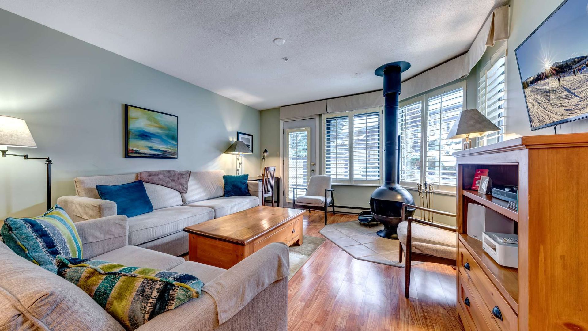 Bright living room with sectional sofa, wood stove, bay windows, and TV on a wooden stand.