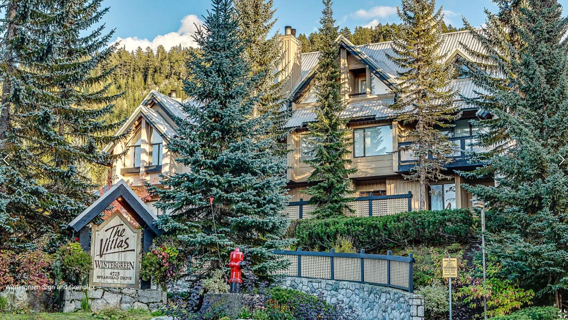 Stone lodge among tall pine trees, with a sign and a person in a red jacket near the entrance