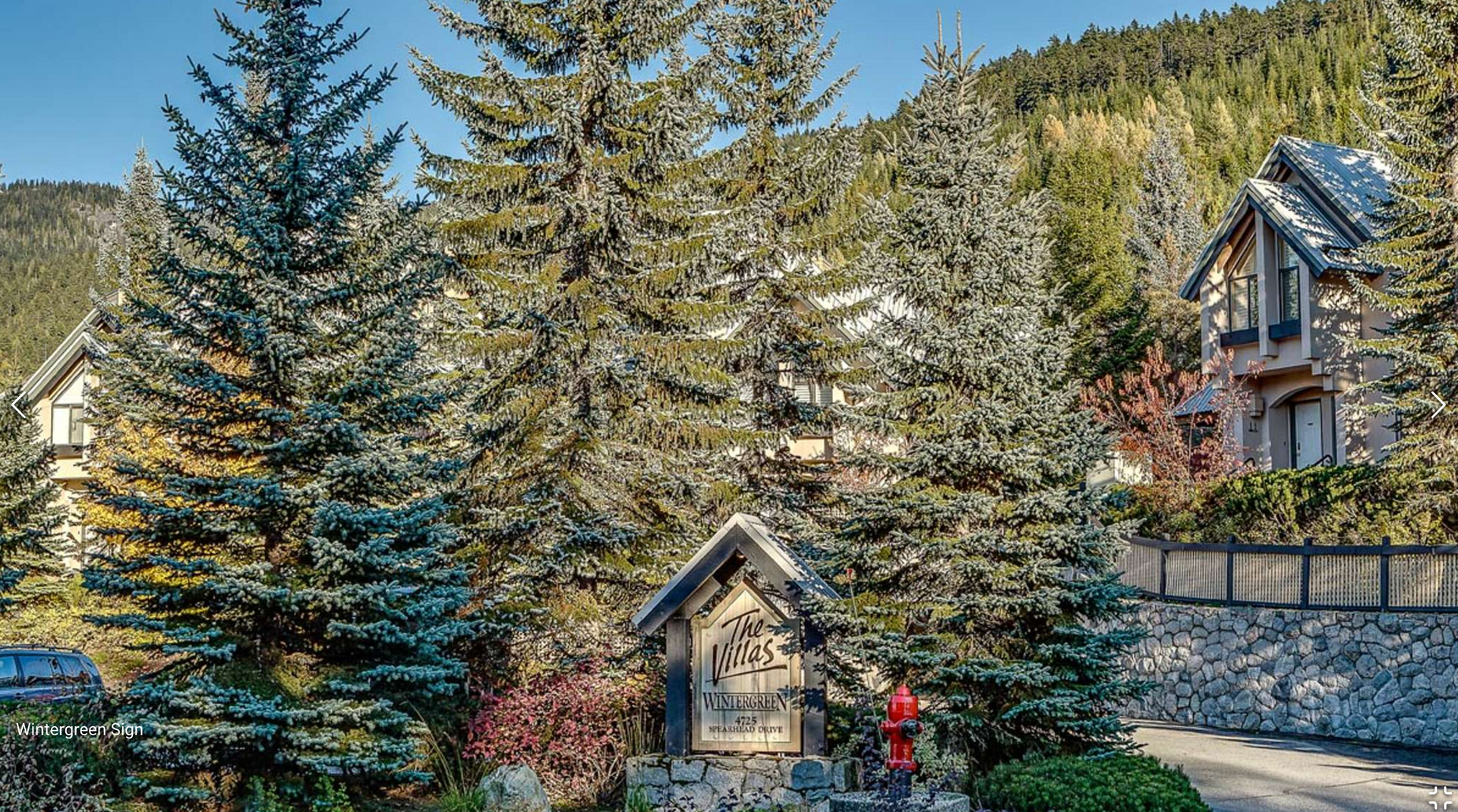 Snowy mountain lodge among tall evergreens, with a small cabin and red-clad person near a stone wall.