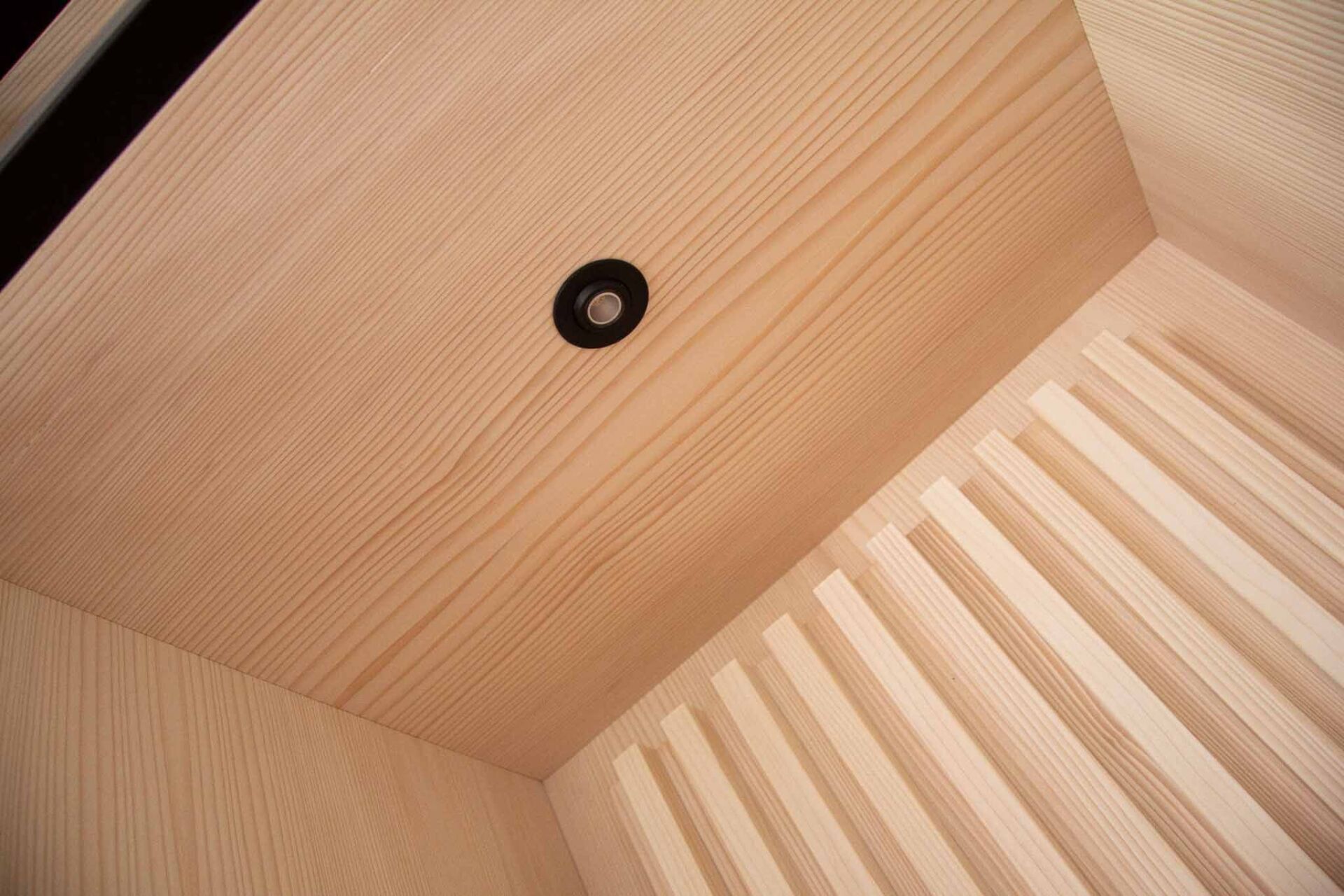 Helle Holzdecke mit schwarzer Rundleuchte und Lamellenwandteil.