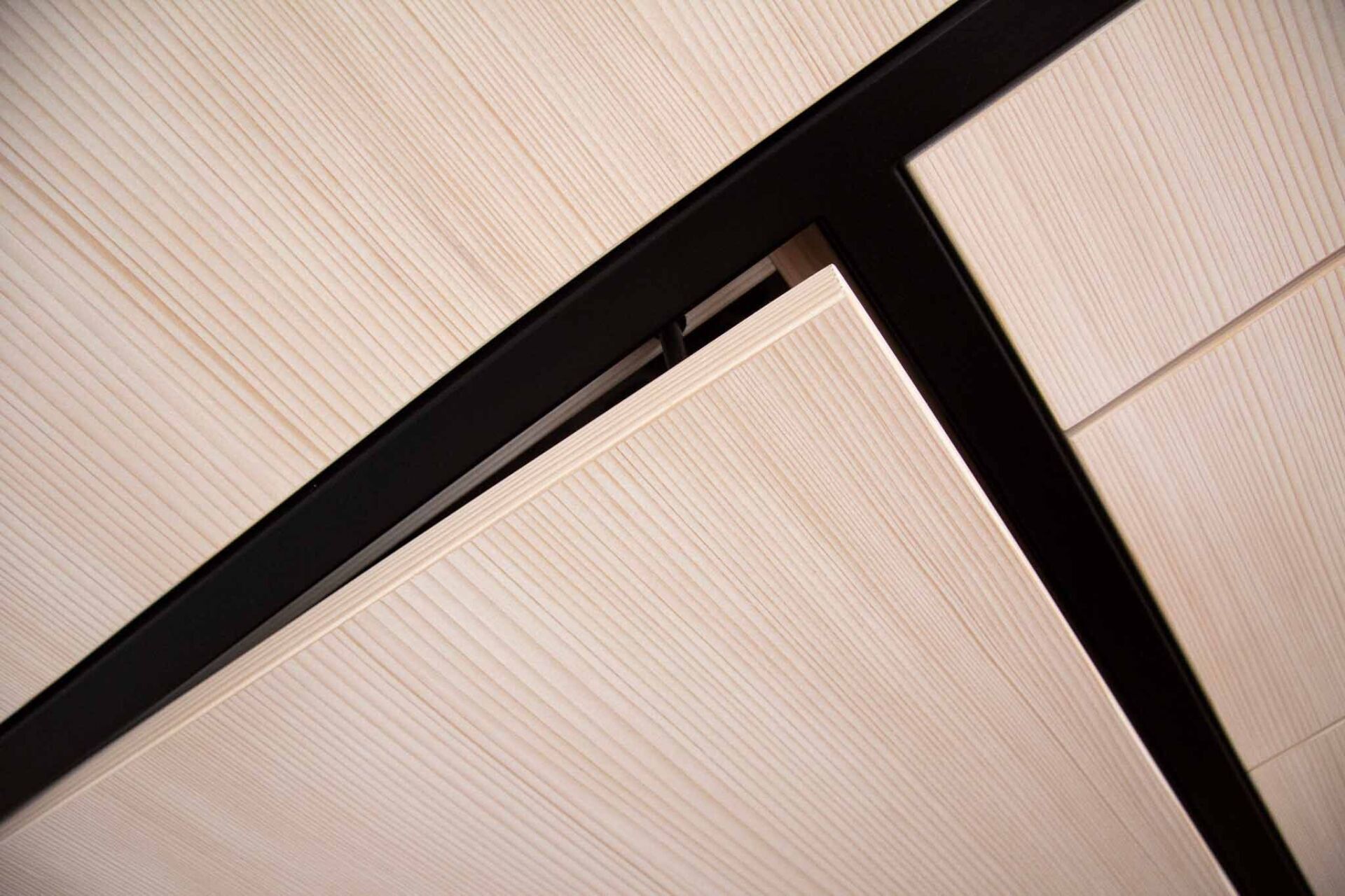 Helle Deckenpaneele mit Holzmaserung und dunklen, schwarzen Strukturbalken in einem abgewinkelten Muster.