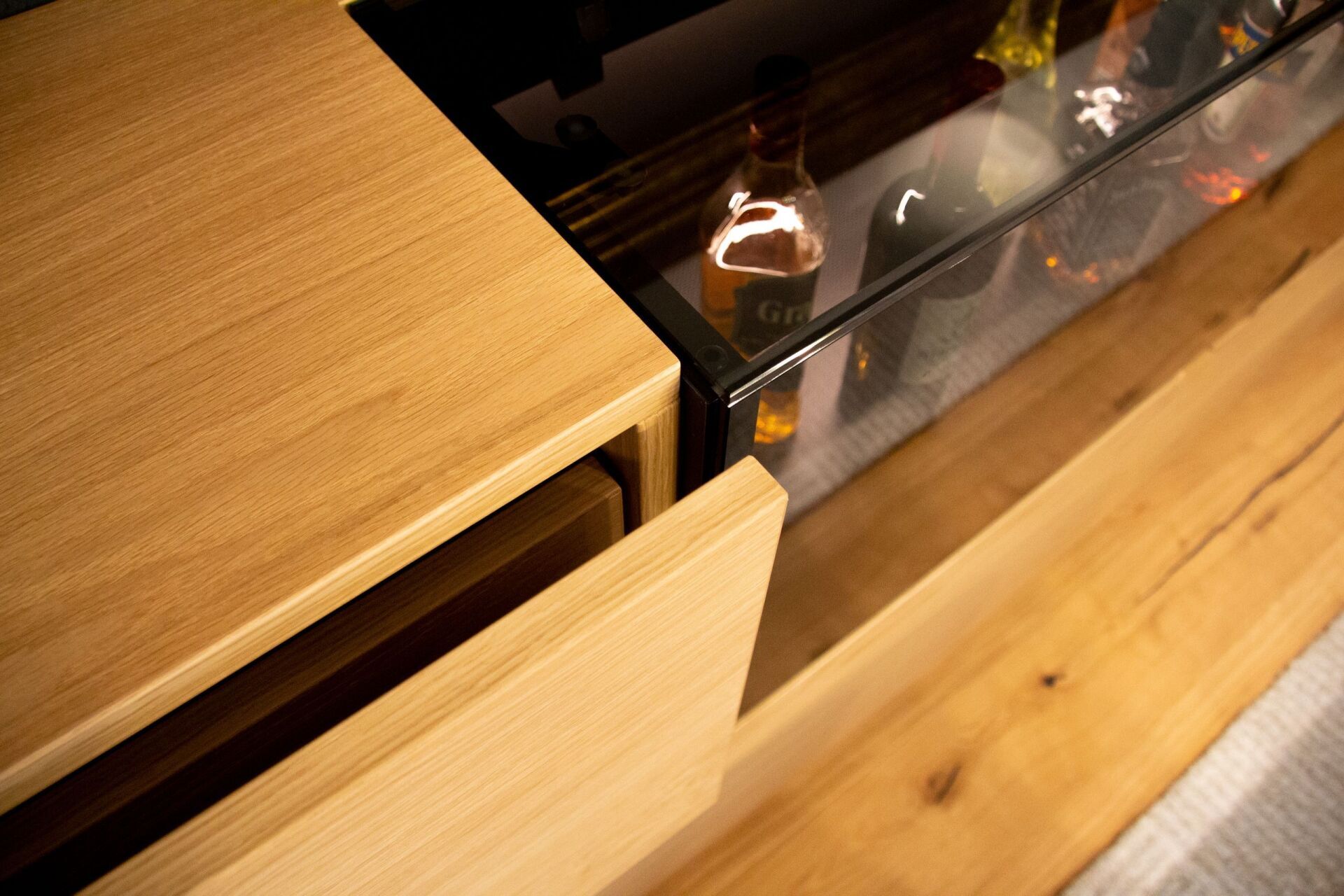 Nahaufnahme eines hellen Holzschranks mit einer offenen Schublade, hinter der sich Glasregale mit Schnapsflaschen befinden.