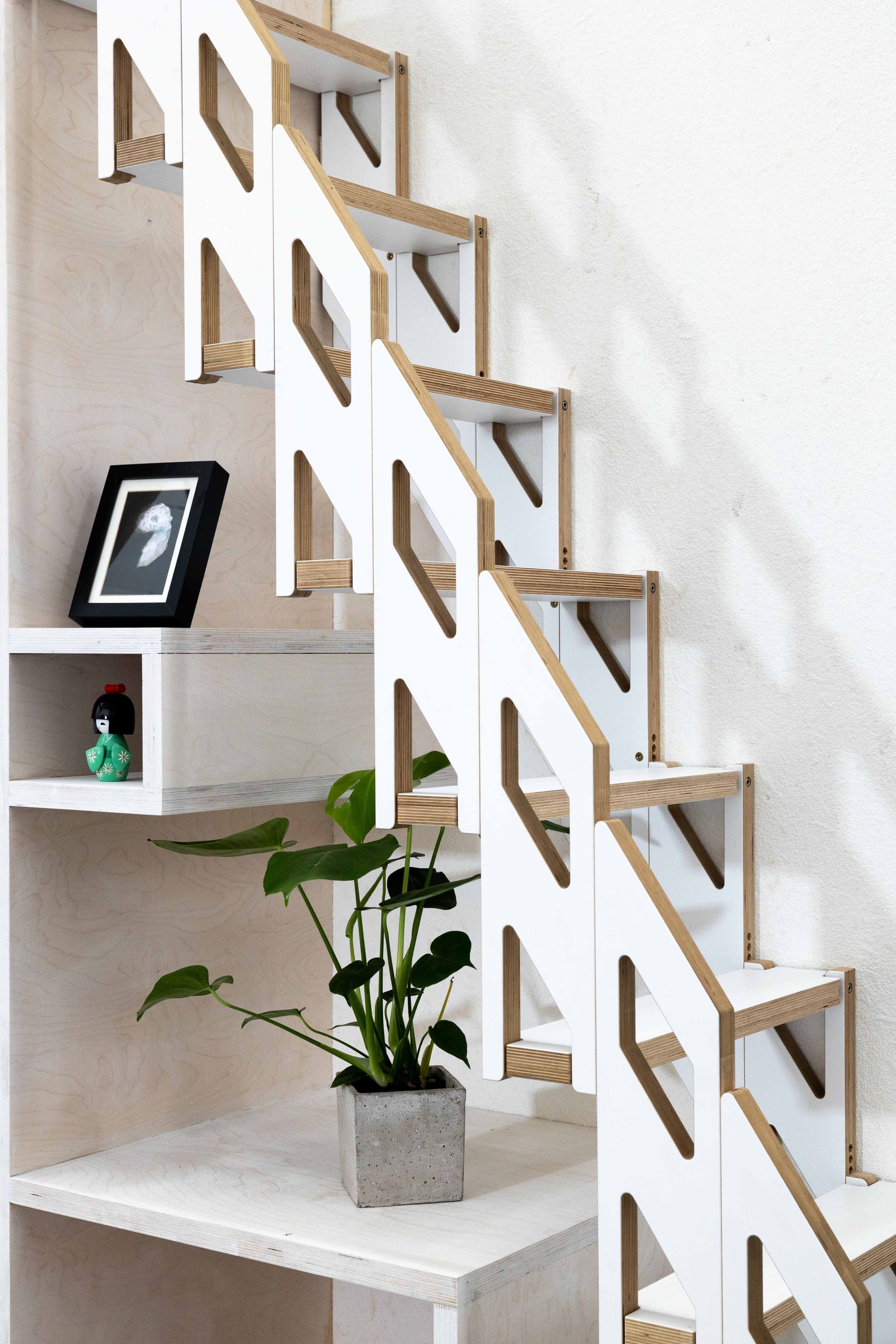 Geometrische Treppe in Weiß und Holztönen mit eingebautem Regal. Unter der Treppe stehen eine Pflanze und ein gerahmtes Foto.