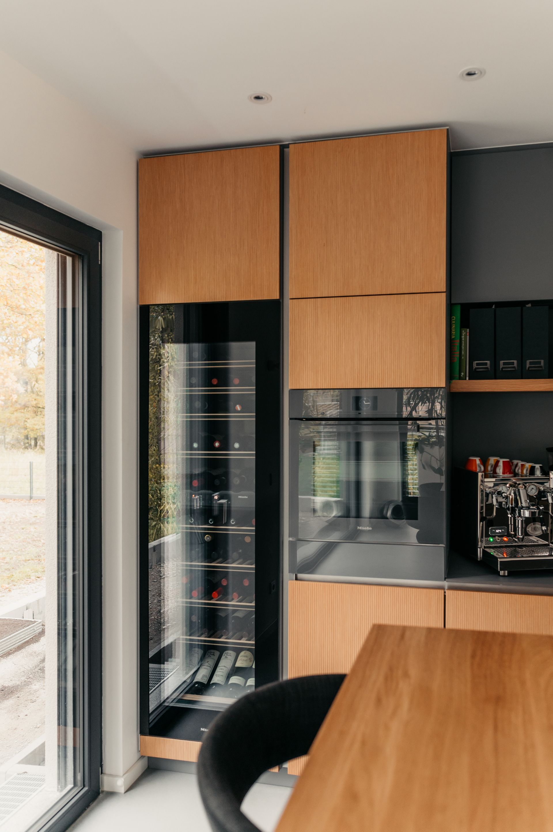 Holzschrank mit eingebautem Weinkühler und Backofen in einer Küche. Schwarze Akzente und ein Tisch sind sichtbar.
