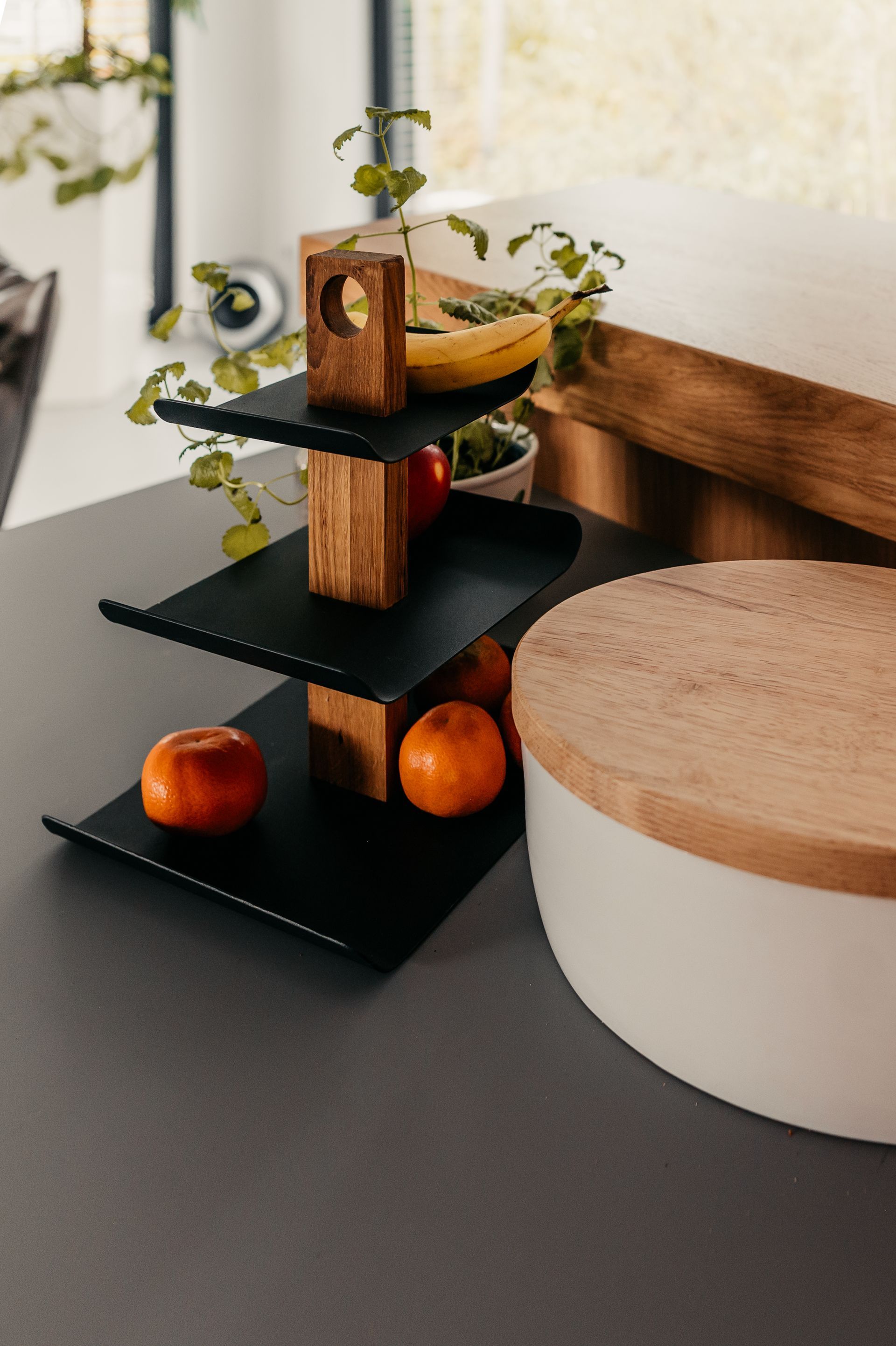 Dreistöckiges schwarzes Tablett mit Obst, Holzstützen und einem weißen Vorratsbehälter mit Holzdeckel auf einer Arbeitsplatte.