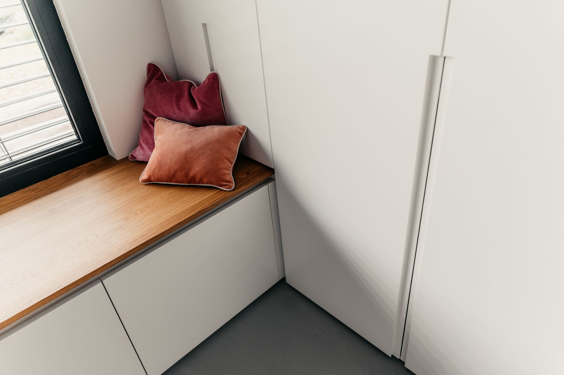 Gemütlicher Fensterplatz mit Holzplatte, weißen Schränken und rosa/orangen Kissen.
