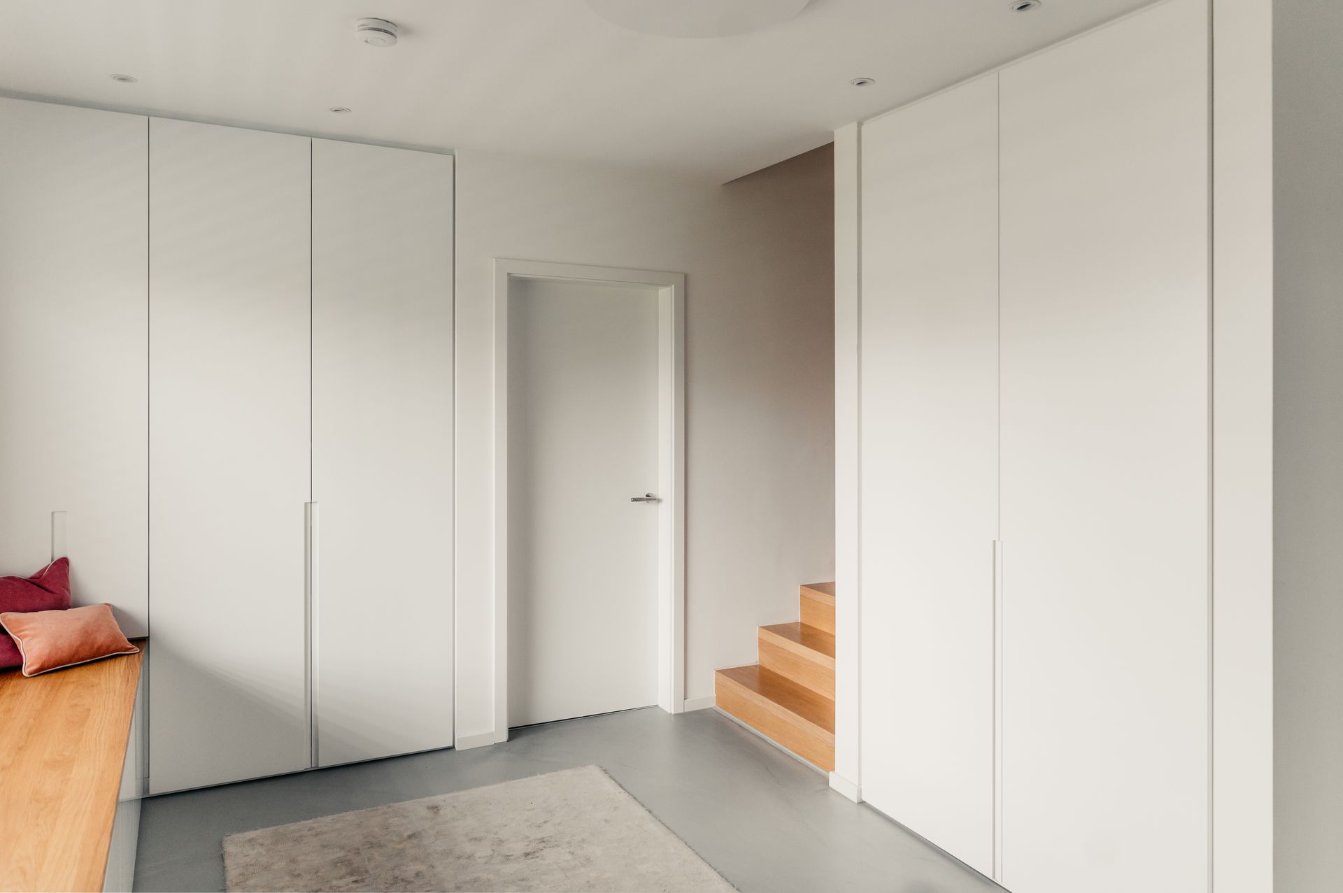 Weißer Raum mit Schränken, einer Tür und einer Treppe nach oben. Ein kleiner Teppich und eine Bank mit Kissen sind sichtbar.