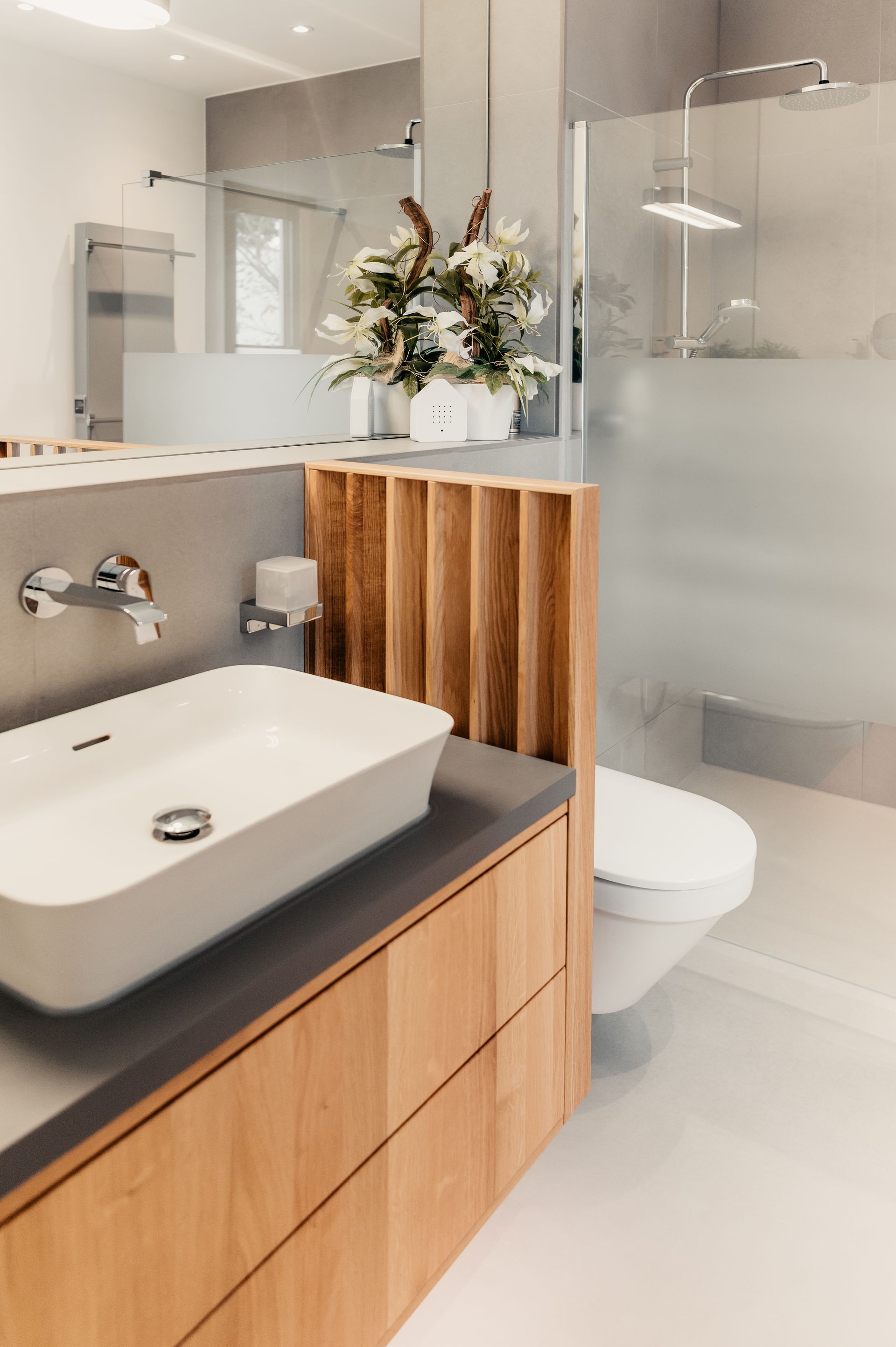 Modernes Badezimmer mit Holzwaschtisch, weißem Waschbecken, Milchglasdusche und Blumen.