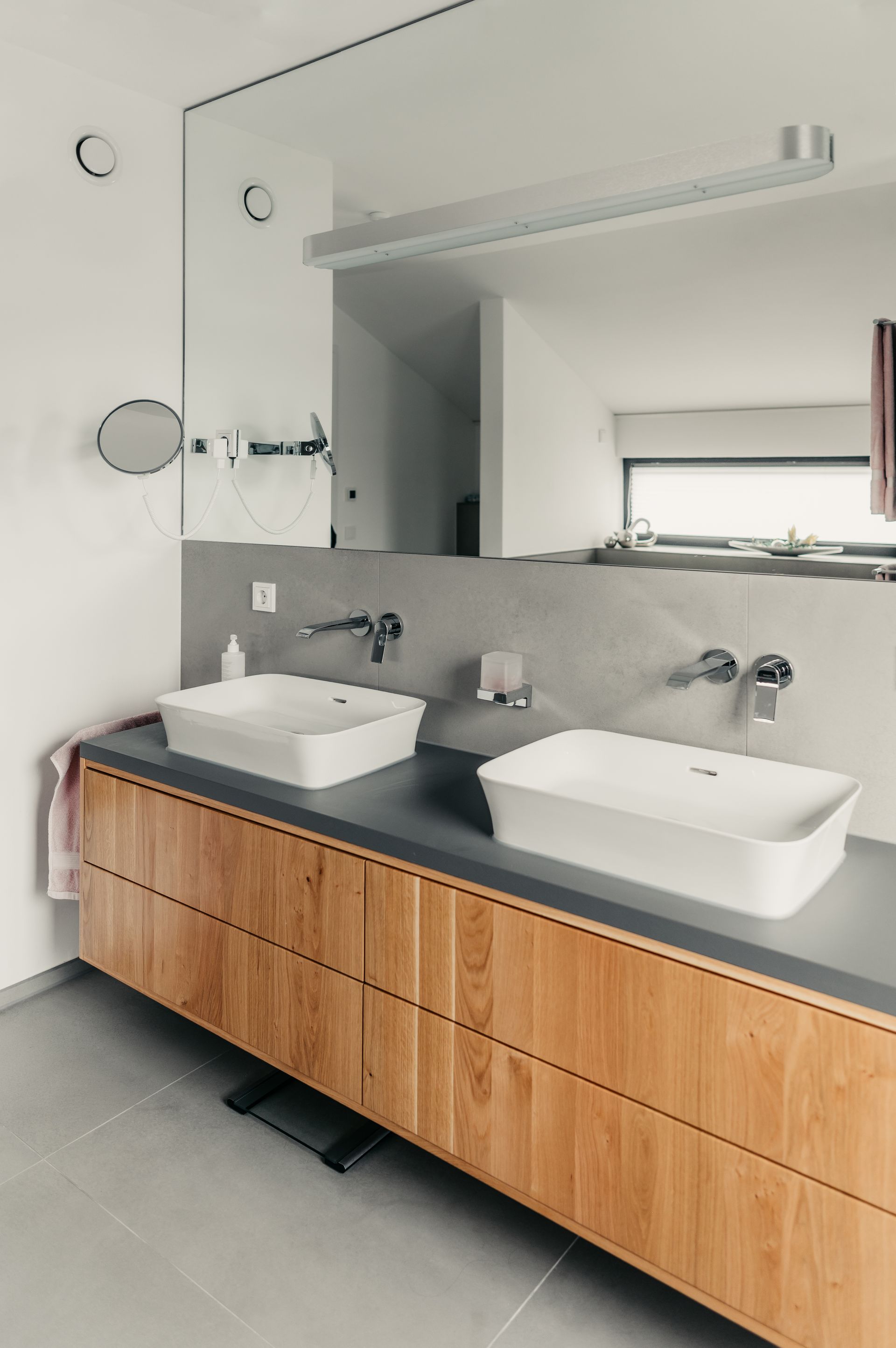 Badezimmer mit zwei weißen Waschbecken auf einem Holzwaschtisch unter einem großen Spiegel. Hellgraue Arbeitsplatte und Wände.