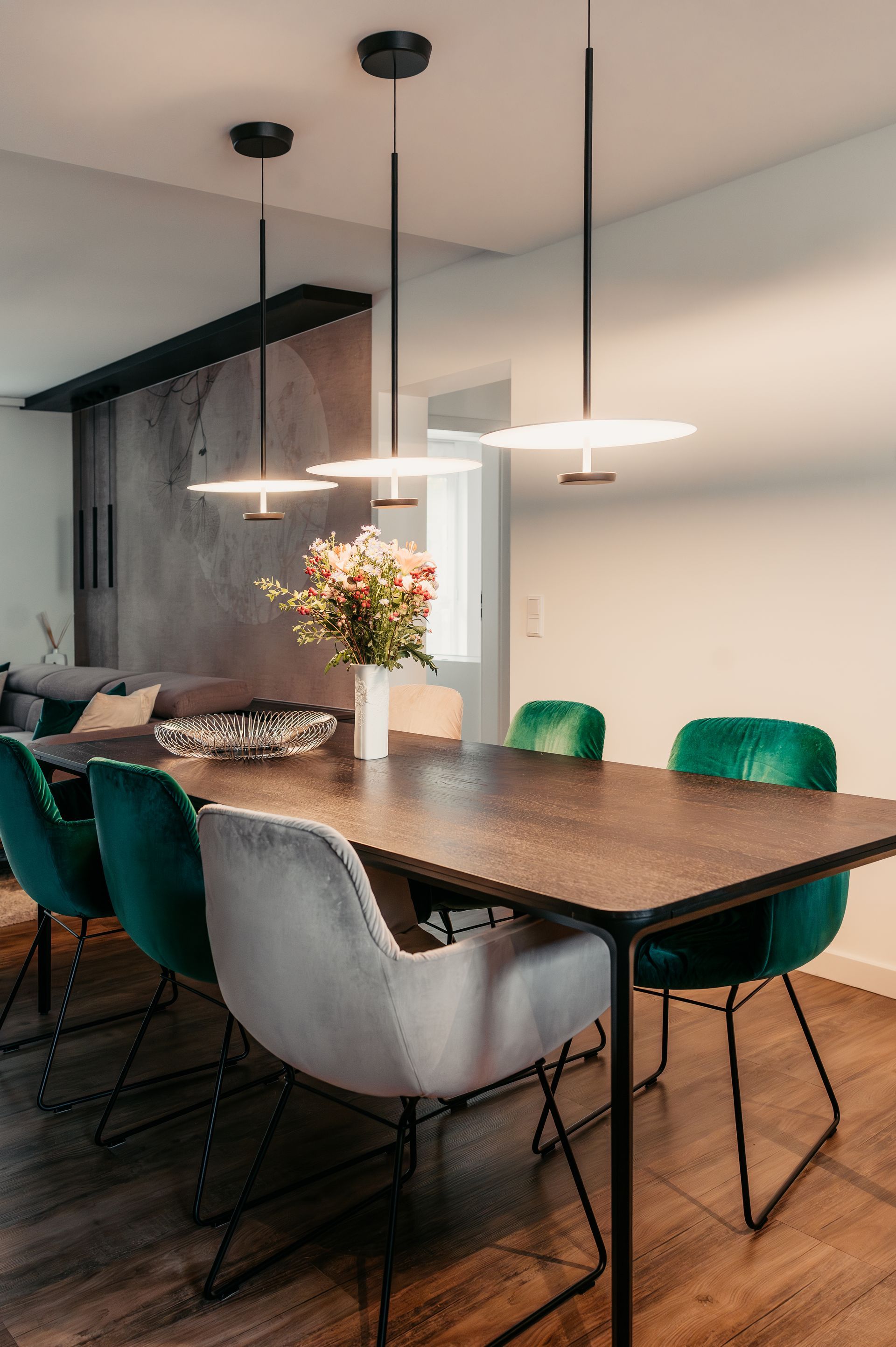 Esszimmer: dunkler Holztisch, grüne Samtstühle, grauer Stuhl, Pendelleuchten, Blumenstrauß, neutrale Wand.