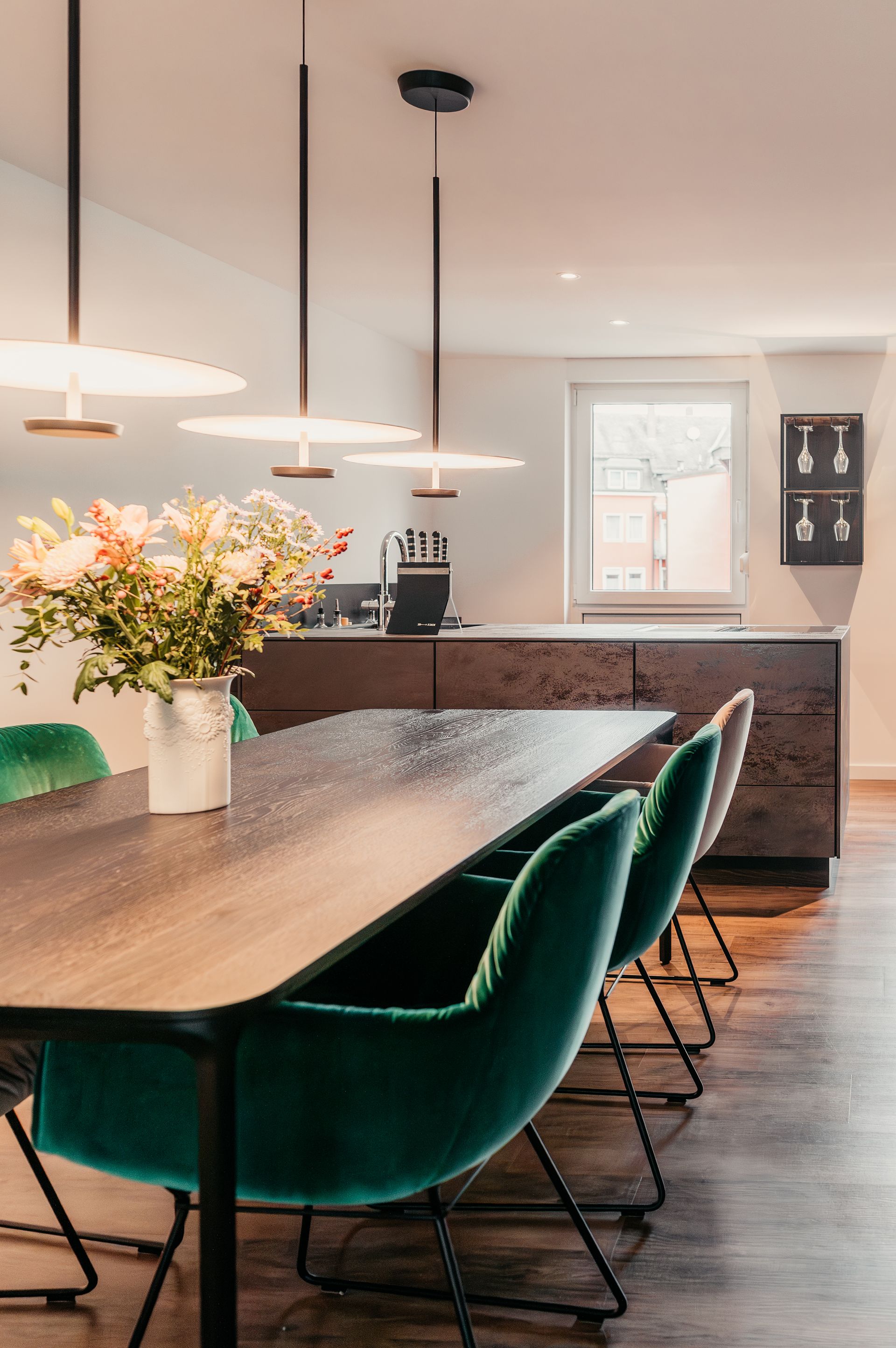 Esszimmer mit langem Holztisch, blaugrünen Samtstühlen, Blumen und Hängelampen.