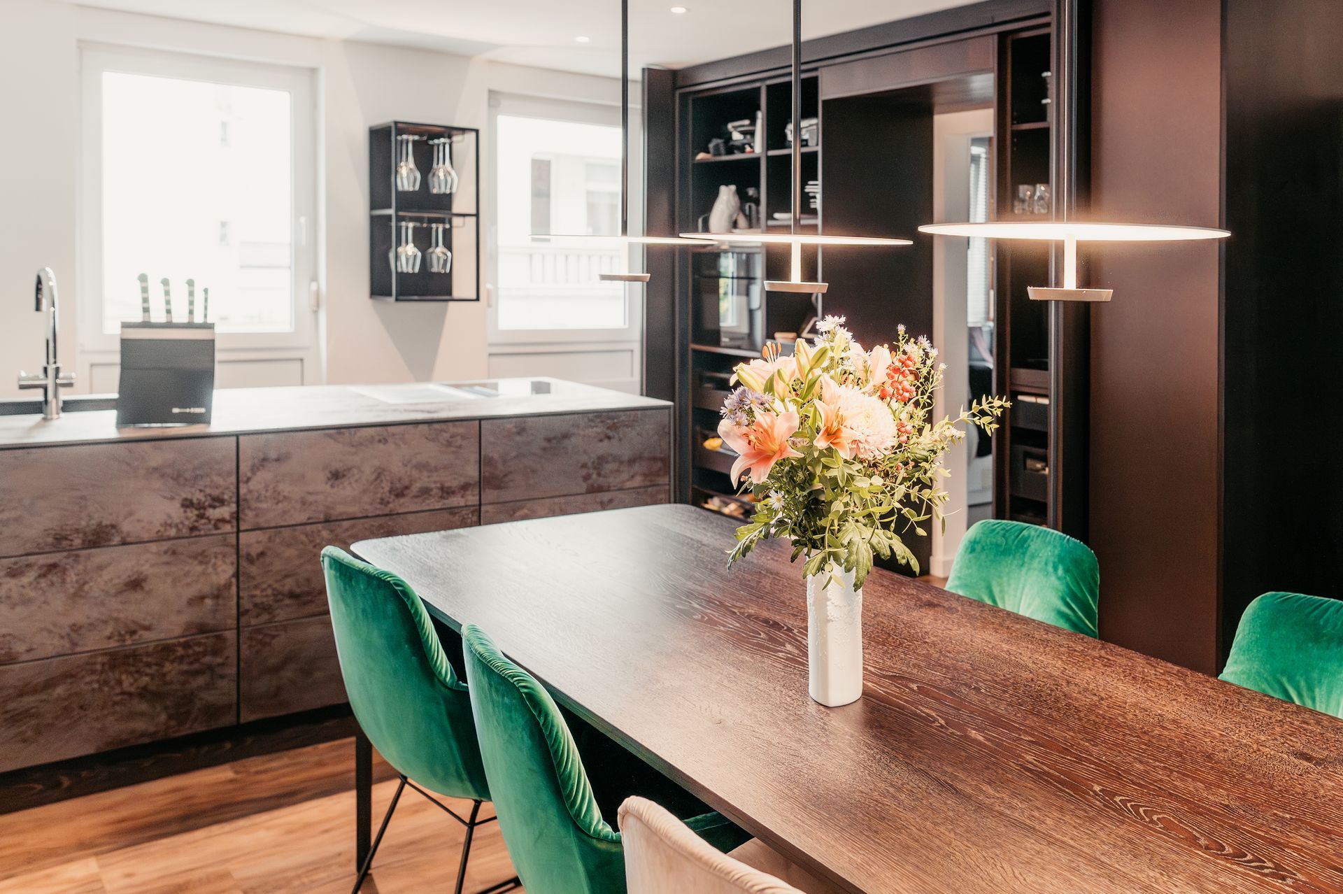 Küche mit Esstisch aus dunklem Holz, smaragdgrünen Stühlen, Steintheke und Blumendekoration.