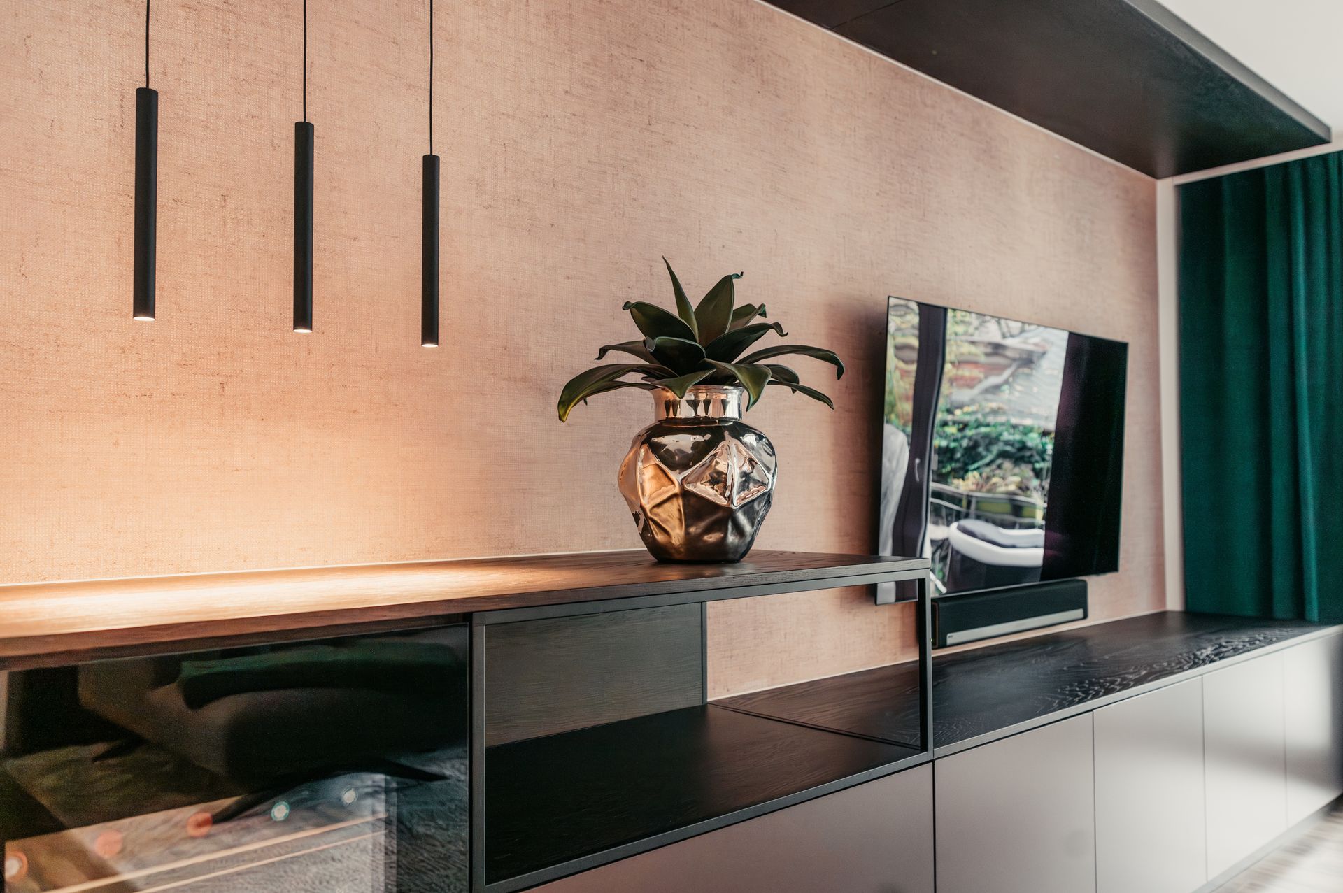 Modernes Wohnzimmer mit Fernseher, dekorativer Vase und Hängelampen vor einer strukturierten pfirsichfarbenen Wand.