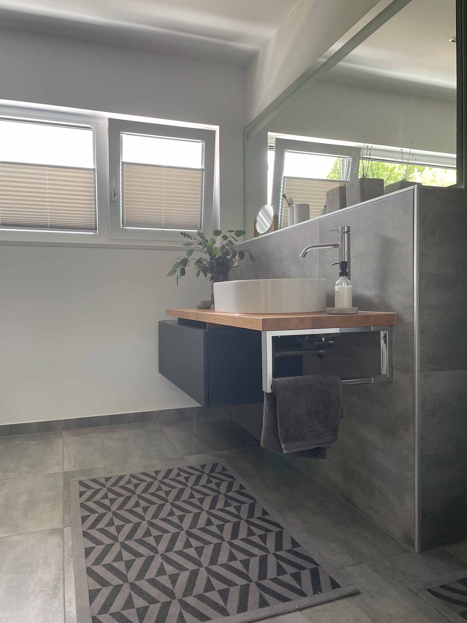 Modernes Badezimmer mit schwebendem Waschtisch, grauen Fliesen, großem Spiegel und geometrischem Teppich.