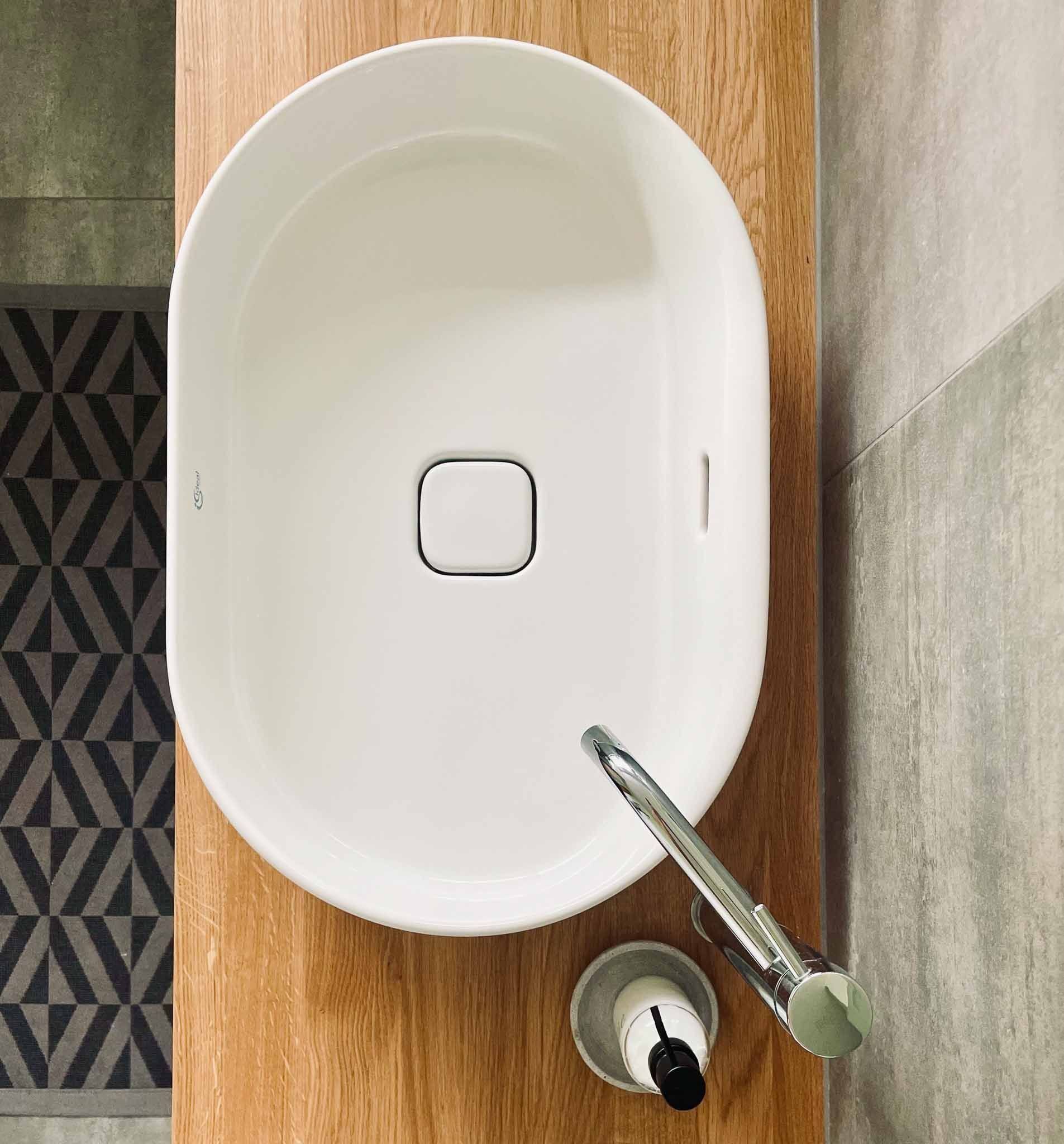 Weißes ovales Waschbecken mit Chromhahn auf einer Holztheke, neben einem gemusterten Teppich und einer grauen Wand.