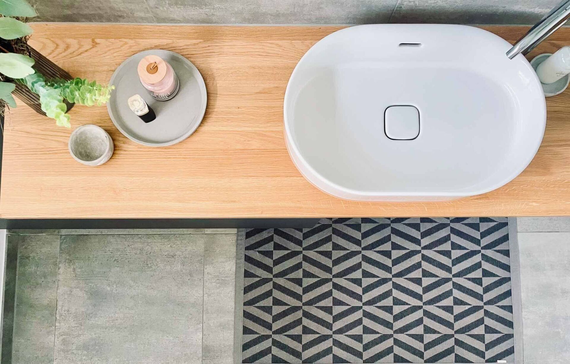 Draufsicht auf ein Waschbecken auf einer Holztheke, mit Zubehör und gemusterten Bodenfliesen.