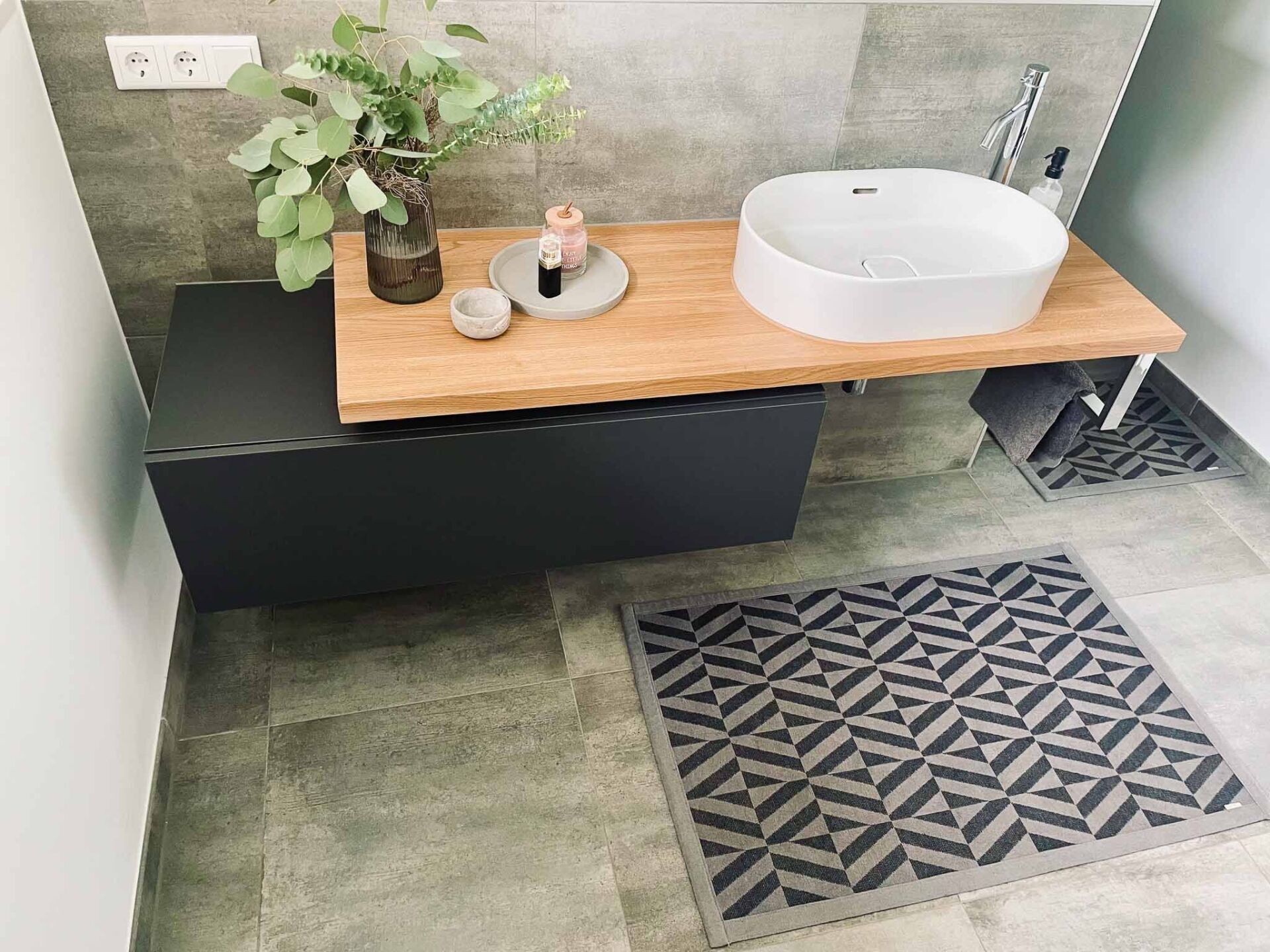 Badezimmer mit grauem Schrank, Holzarbeitsplatte, weißem Waschbecken und grau gemustertem Teppich.