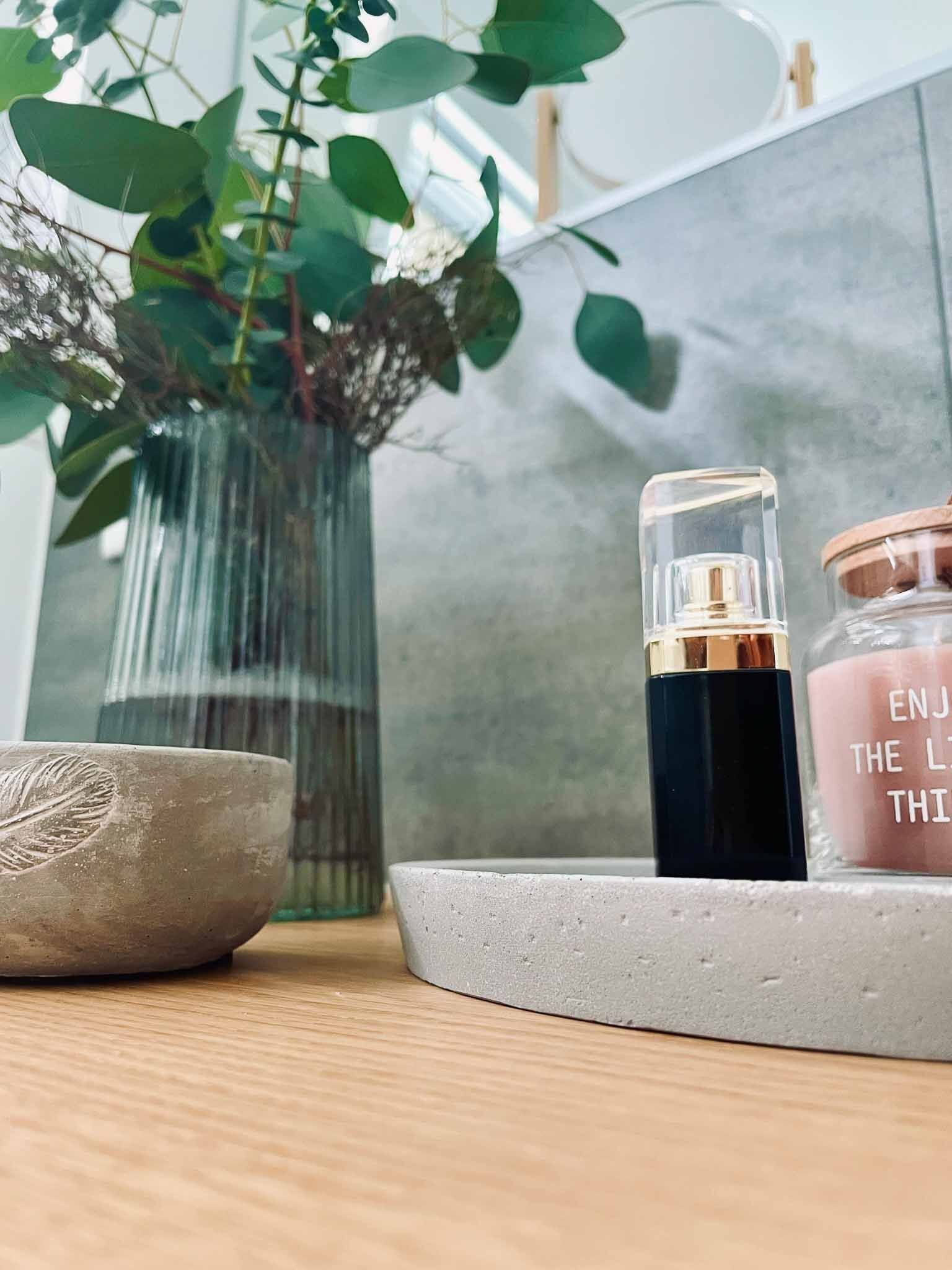 Eine Badezimmerablage mit einer Vase mit Eukalyptus, Hautpflegeprodukten und einer kleinen Schüssel, alles in einer neutralen Farbpalette.