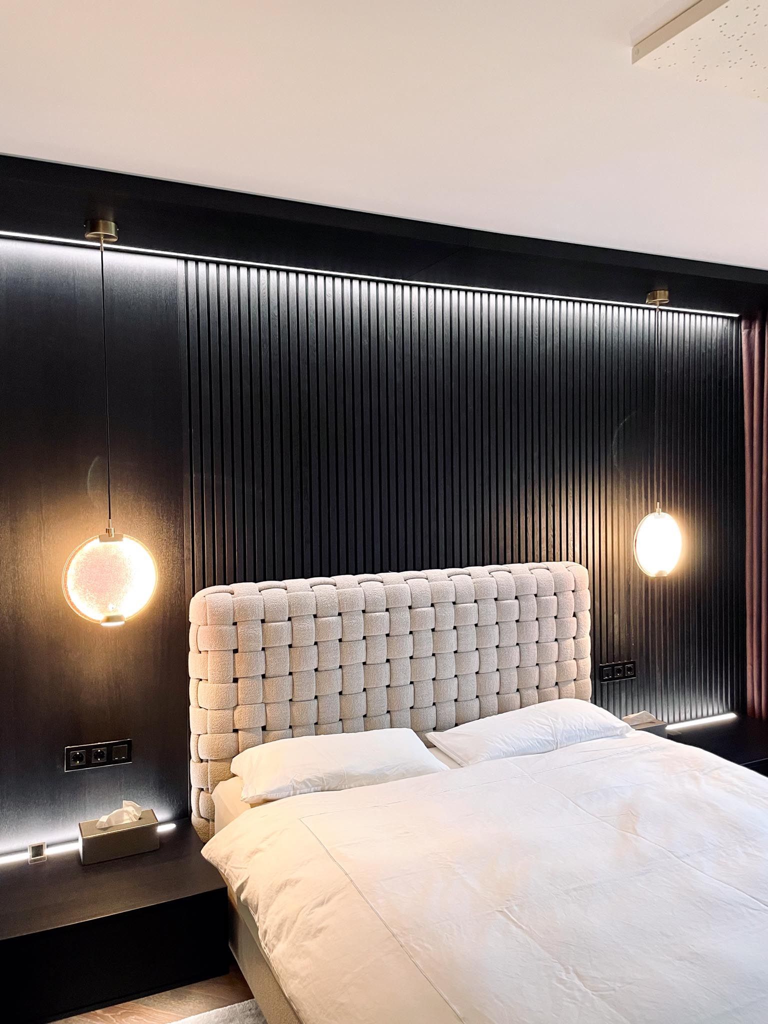 Schlafzimmer mit schwarzer Akzentwand, gewebtem Kopfteil, kugelförmigen Leuchten und Bett mit weißer Bettwäsche.