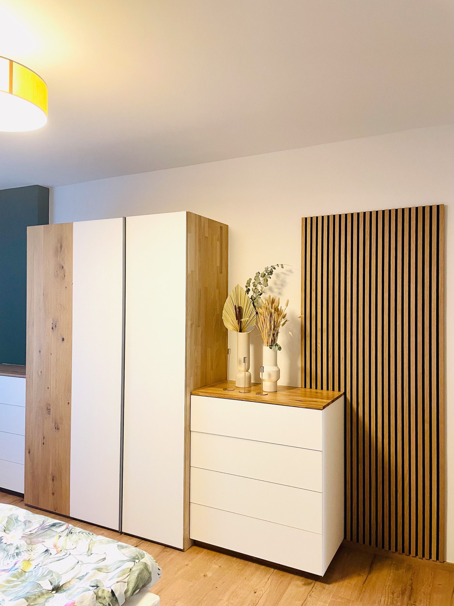 Schlafzimmer mit weißem Schrank, Kommode und Akzentwand aus Holz. Blumen auf der Kommode.