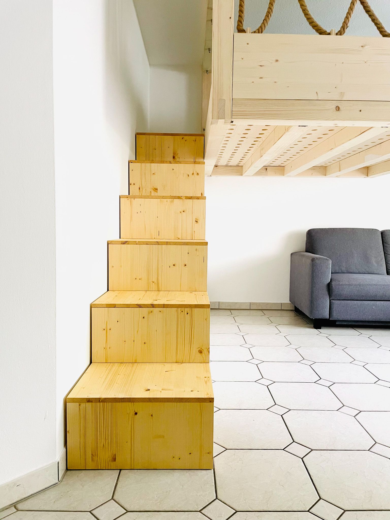 Holztreppe, die zu einem Loft führt, weiße Wände, graues Sofa und gemusterter Boden.