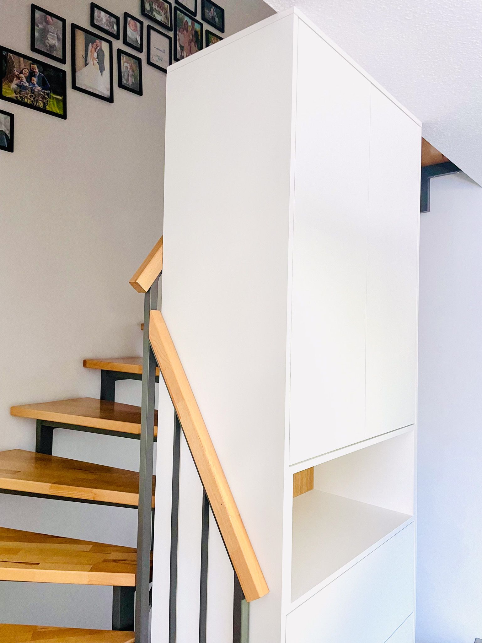 Treppe mit Holzstufen und Metallgeländer, daneben ein weißer Lagerschrank. Schwarz gerahmte Fotos an der Wand.