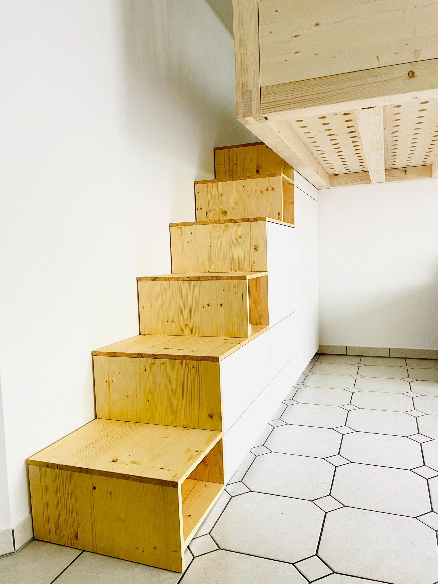 Eine Holztreppe an einer weißen Wand führt zu einem Loft. Sechseckiger Fliesenboden.
