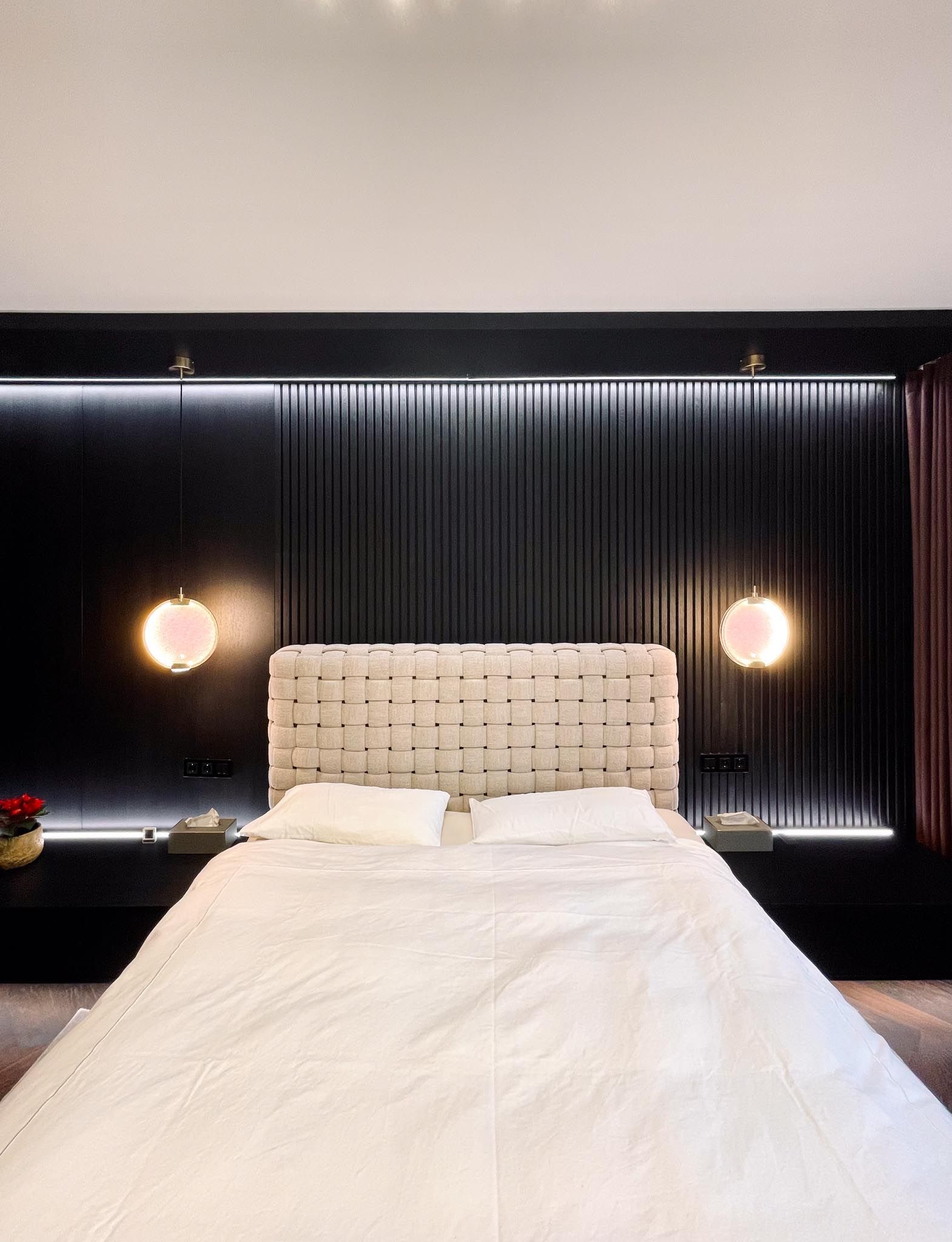 Gemütliches Schlafzimmer mit geflochtenem Kopfteil, schwarzer Wandverkleidung und zwei kugelförmigen rosa Lampen.