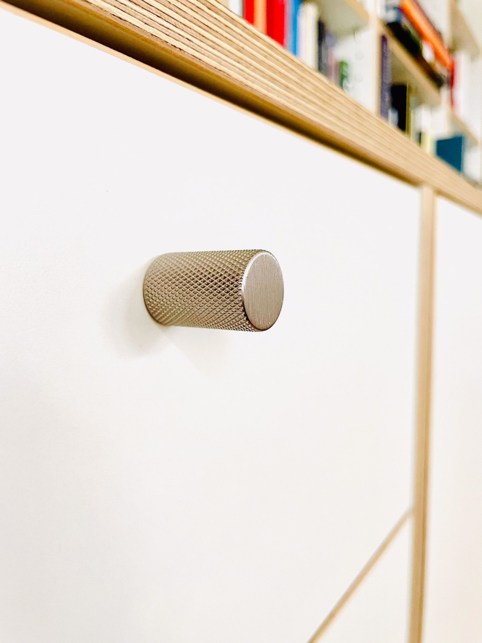 Nahaufnahme eines weißen Schranks mit strukturiertem silbernem Griff. Holzverkleidung und ein teilweise sichtbares Bücherregal im Hintergrund.