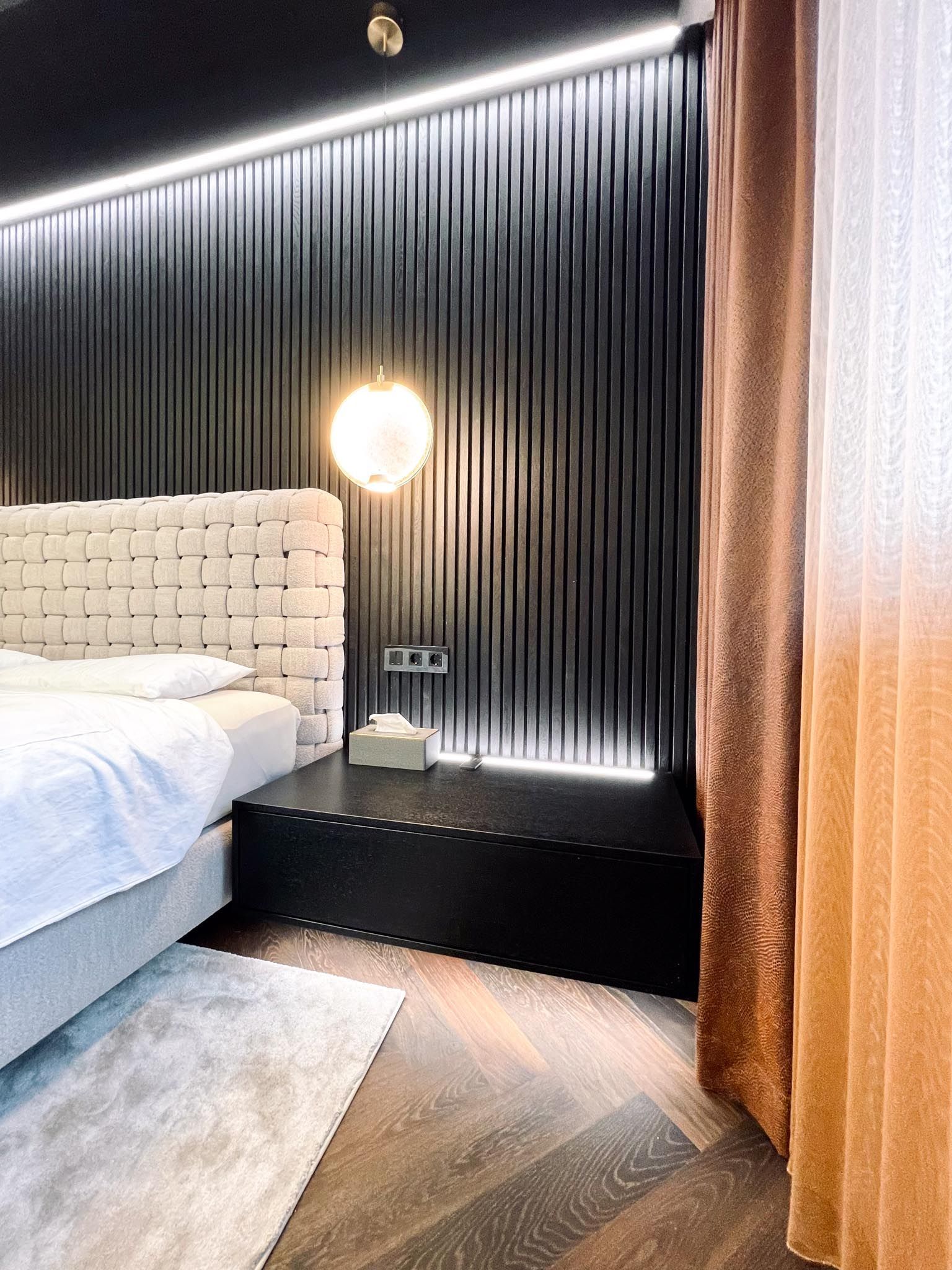 Schlafzimmer mit schwarzer gerippter Wand, gewebtem Kopfteil, runder Lampe und braun-pfirsichfarbenen Vorhängen.