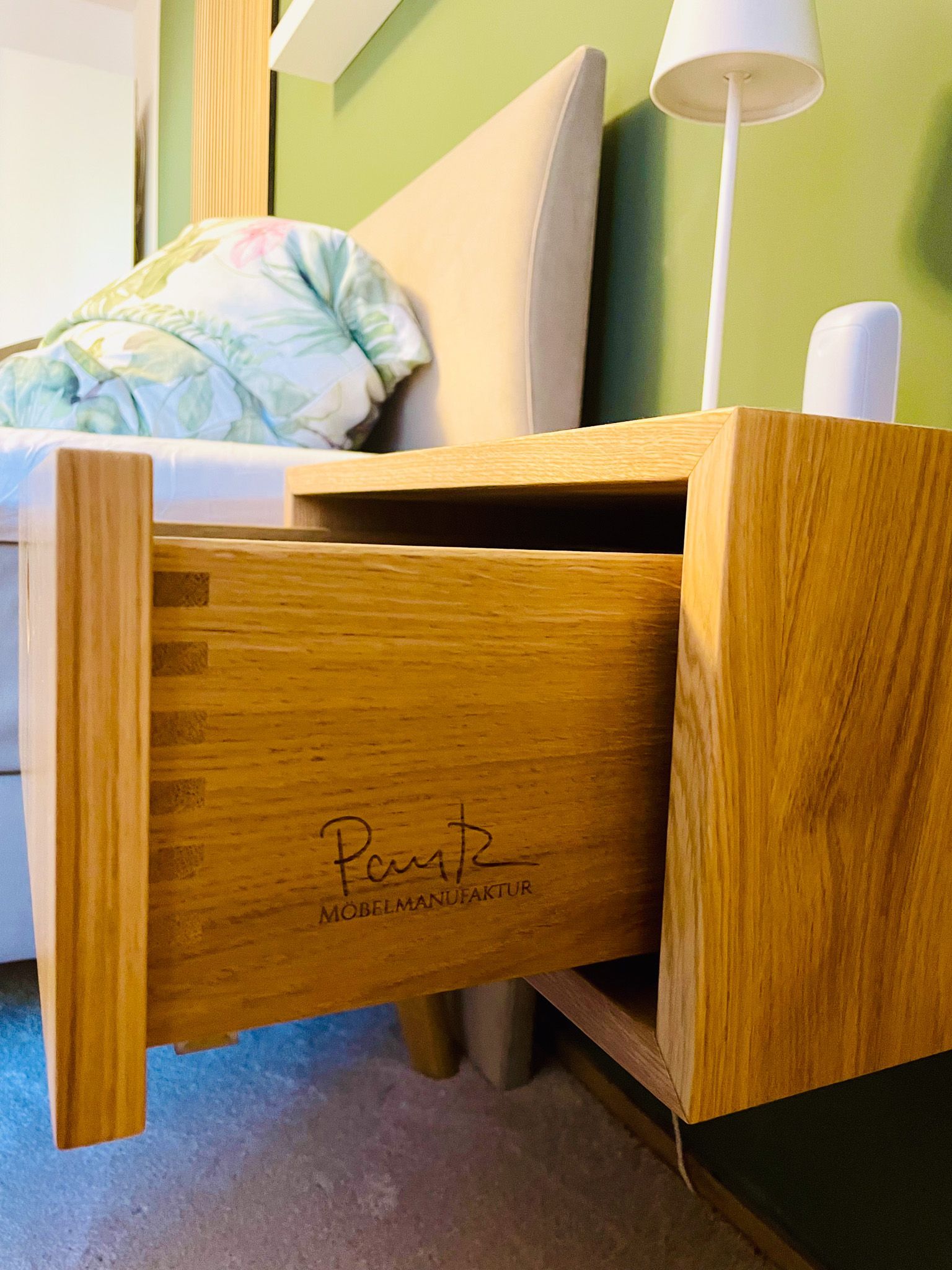 Nachttisch aus Holz mit offener Schublade, Schwalbenschwanzverbindung, Lampe und Bett mit geblümtem Kissen.