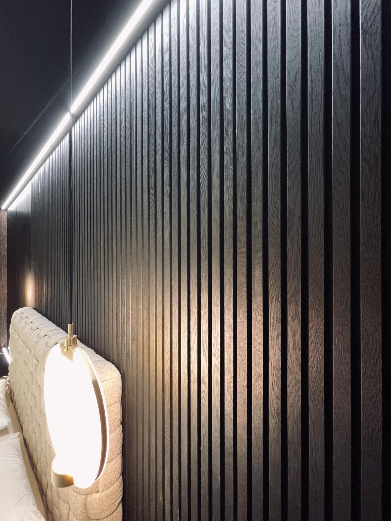 Dunkle Holzlattenwand mit einer modernen Leuchte und einem Teil eines beigen Kopfteils.