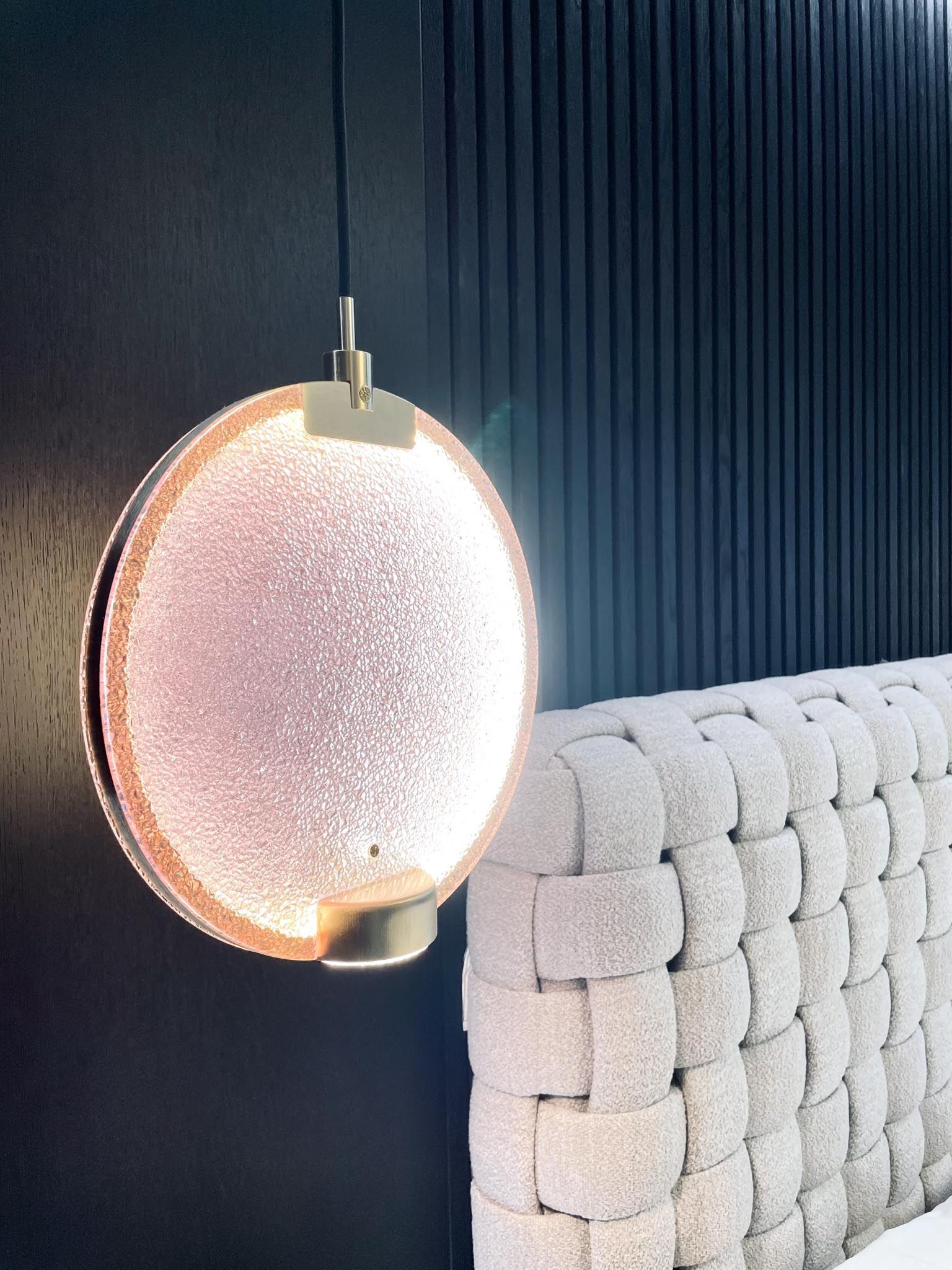 Modernes Schlafzimmer mit runder, beleuchteter Pendelleuchte und geflochtenem Kopfteil. Dunkle Wand im Hintergrund.