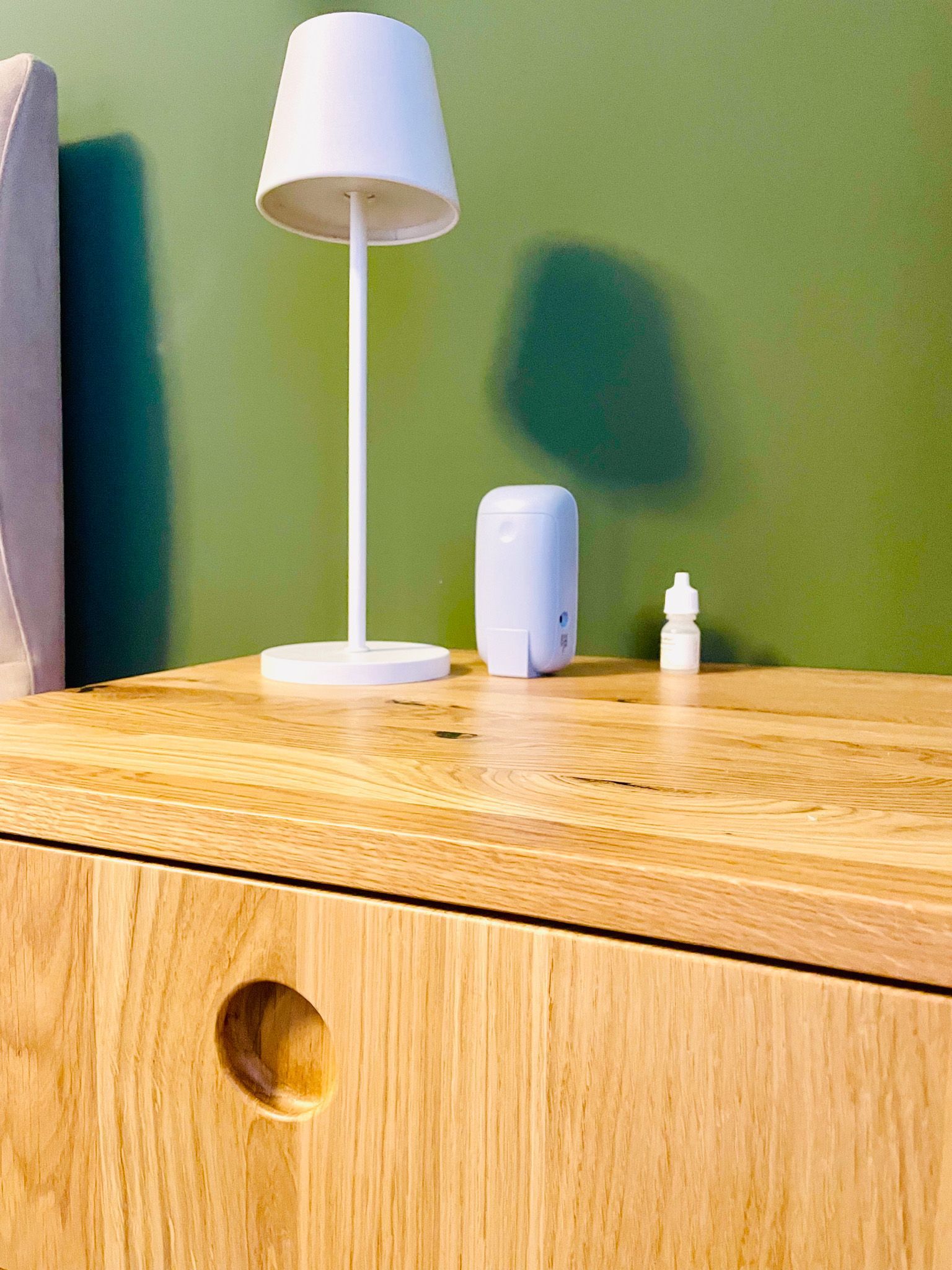 Weiße Lampe, Luftreiniger und Augentropfen auf einem hölzernen Nachttisch vor einer grünen Wand.
