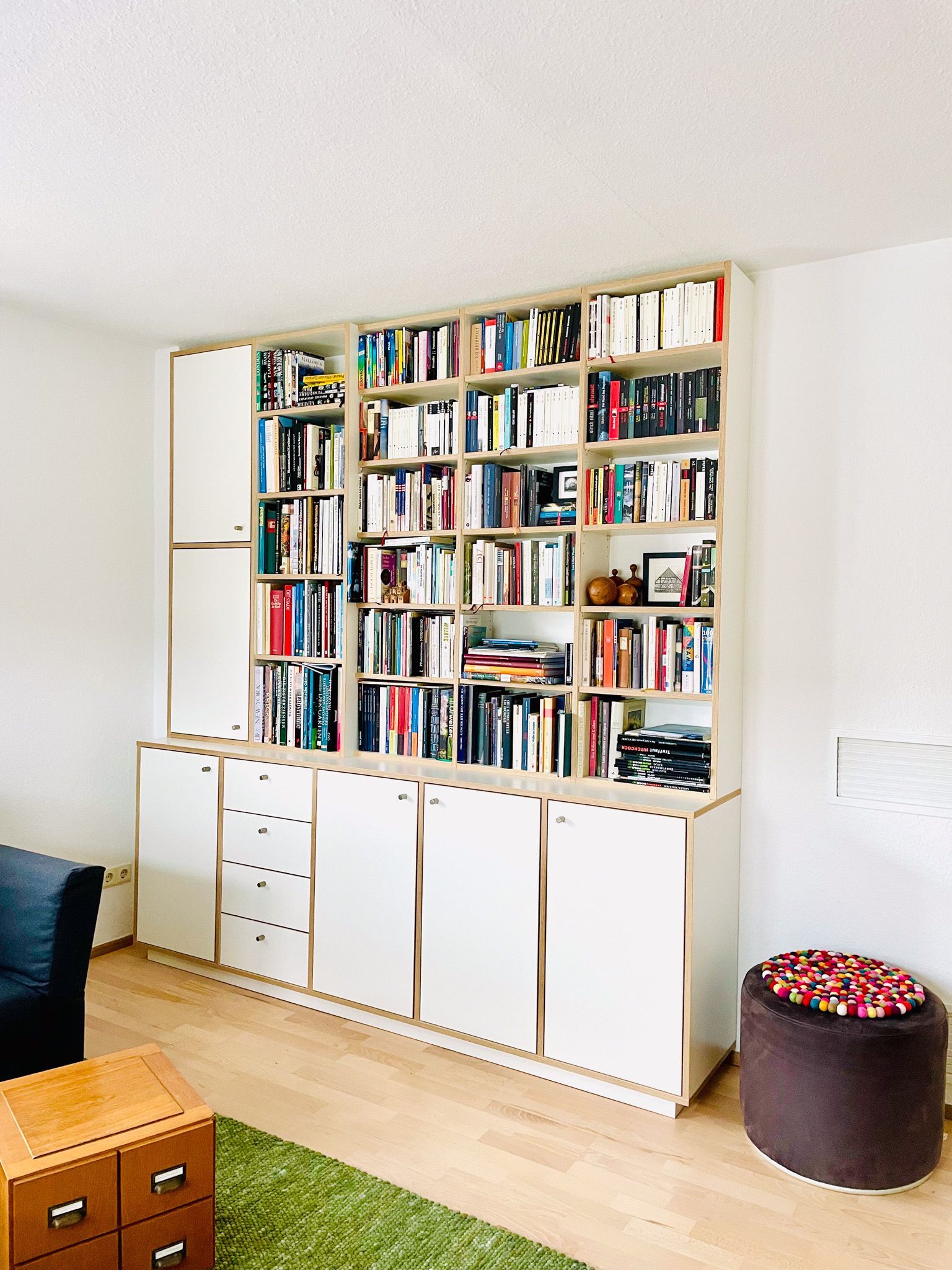 Weißes eingebautes Bücherregal voller Bücher und Lagerschränke. Der Raum verfügt über Holzböden, einen grünen Teppich und eine braune Ottomane.