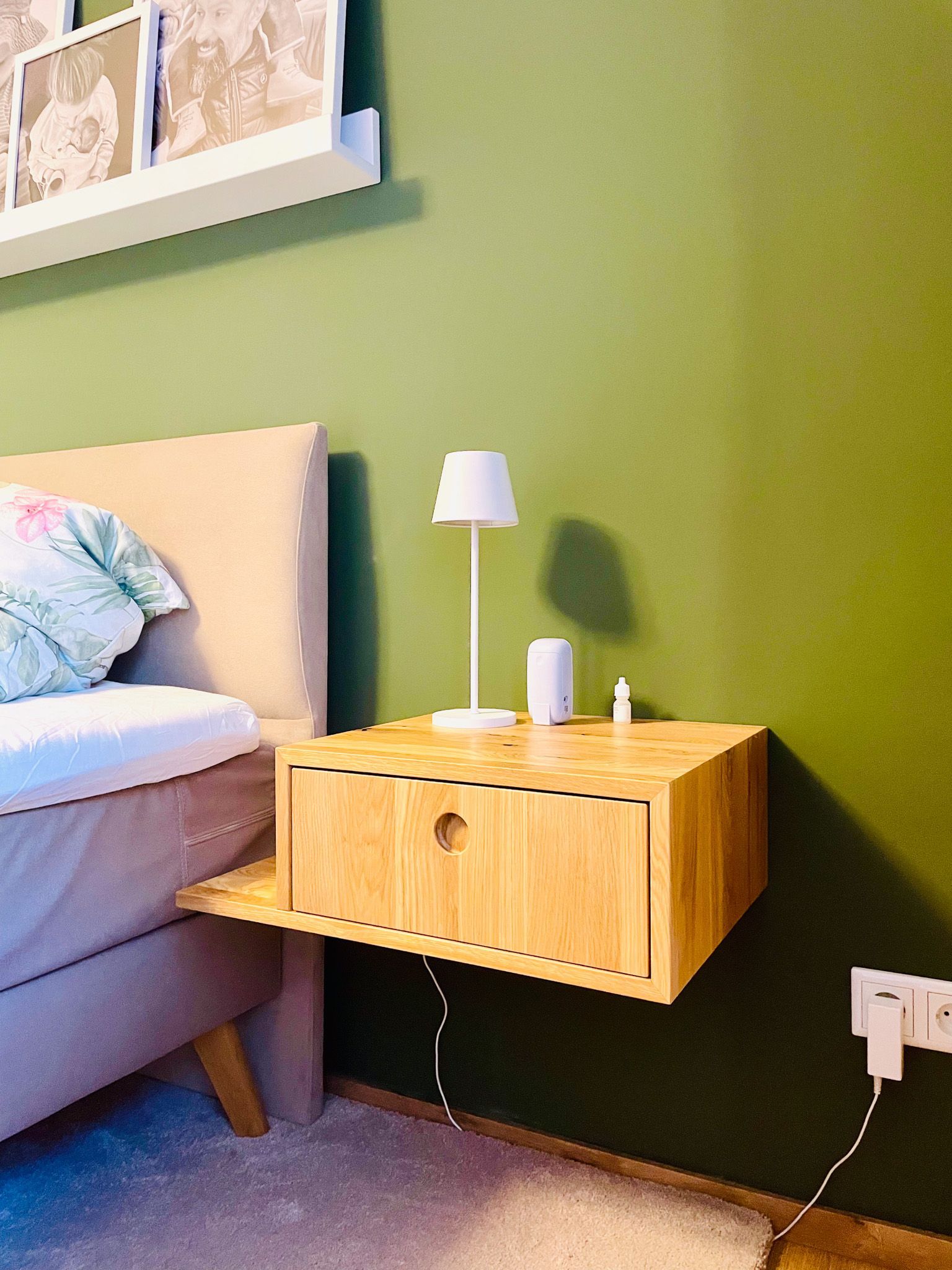 Schwebender Nachttisch aus Holz neben dem Bett, mit Lampe, an grüner Wand.