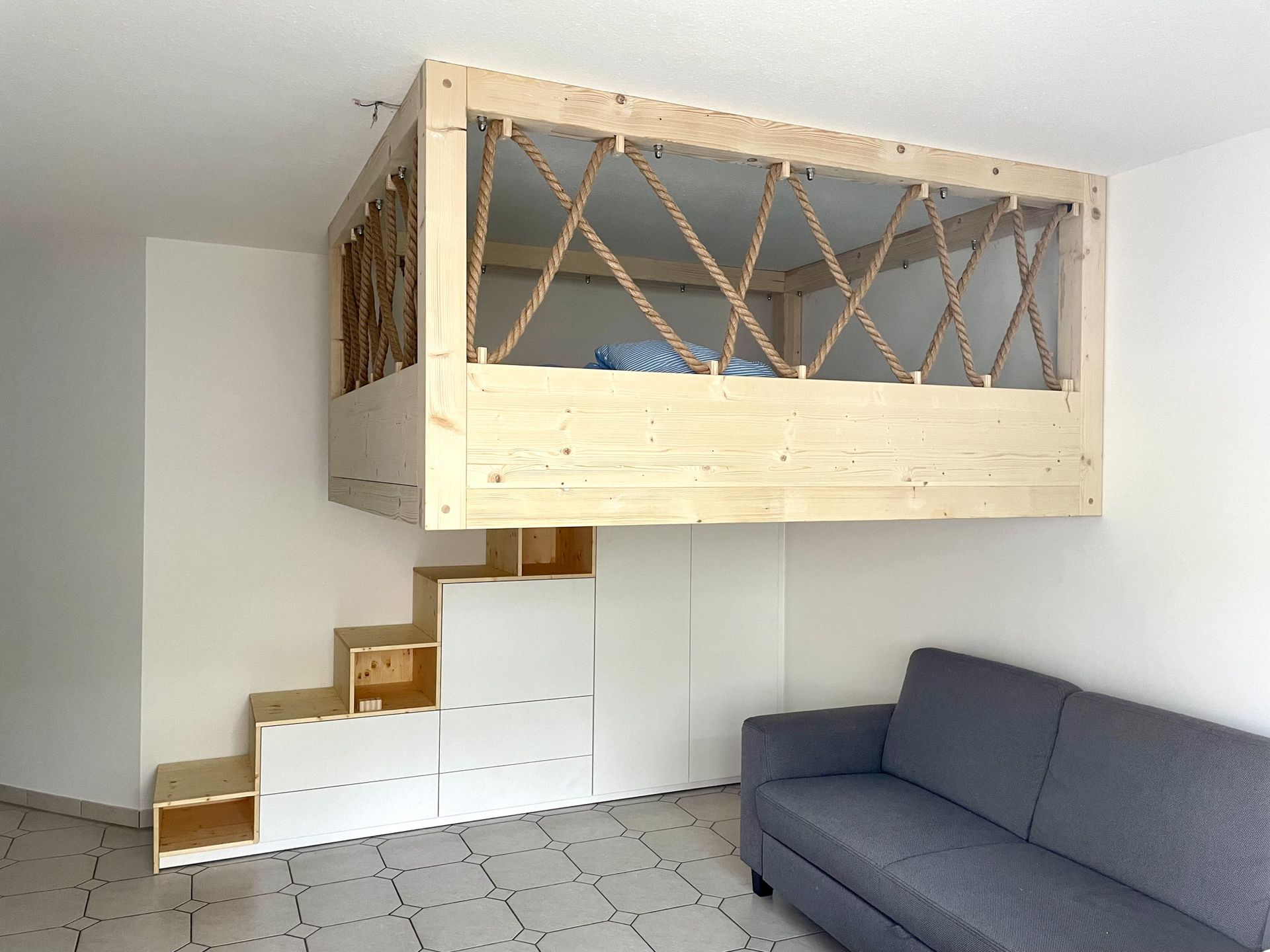 Hochbett mit Seilgeländer und eingebauter Treppe über einem Sofa in einem weißen Raum.