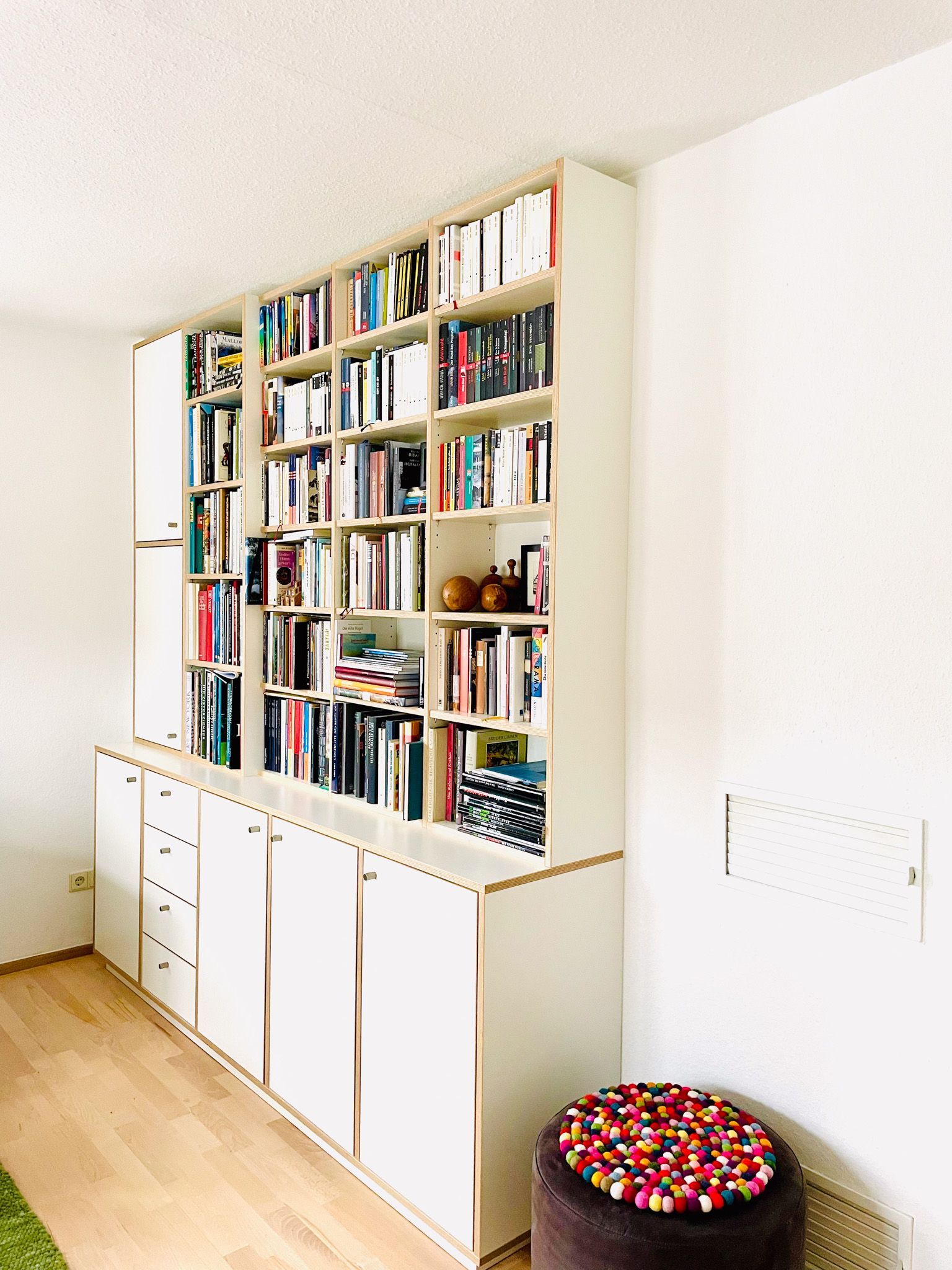 Eingebautes weißes Bücherregal voller Bücher vor einer weißen Wand in einem hellen Raum; davor ein kleiner Hocker.