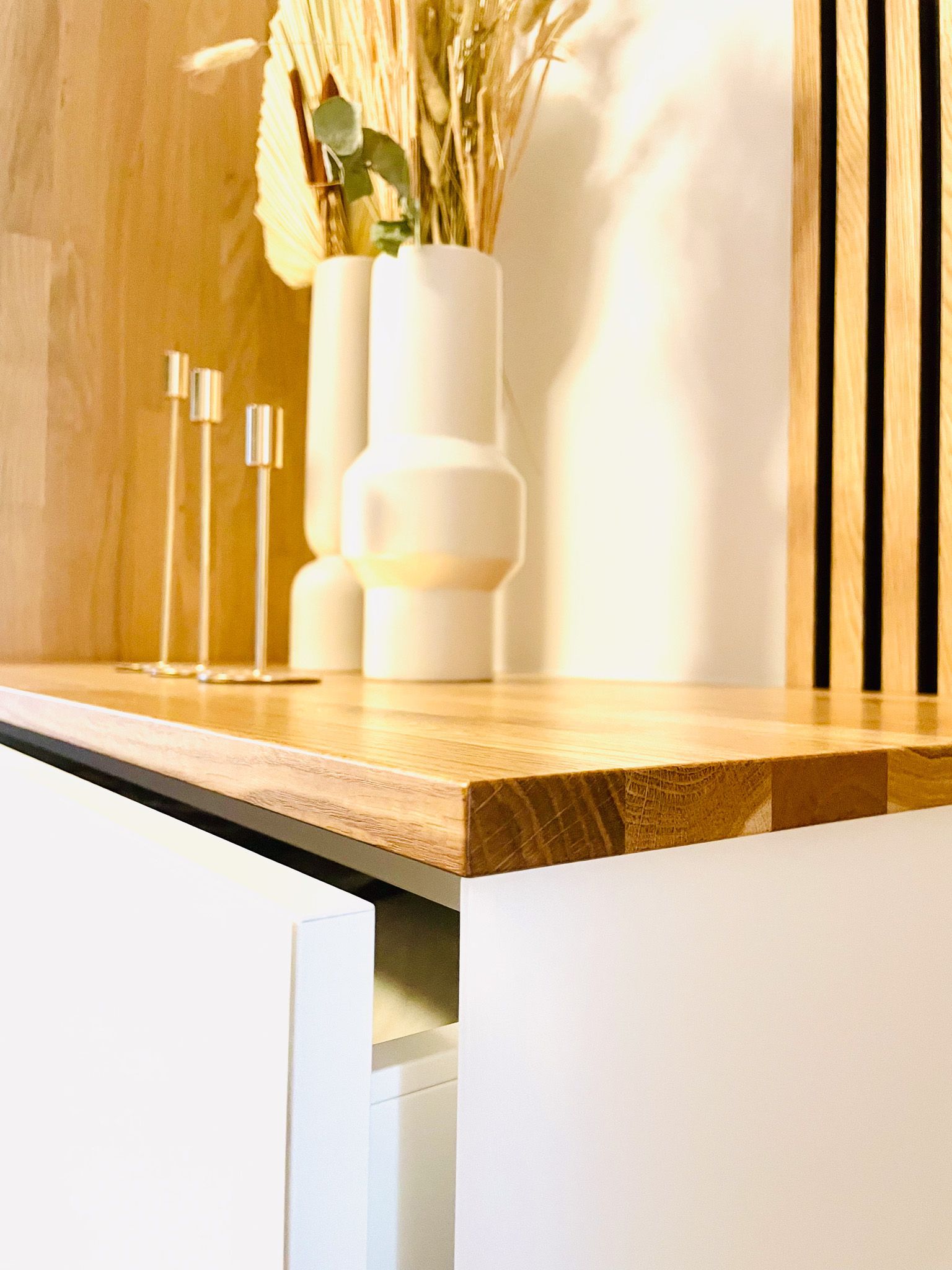 Holzarbeitsplatte mit weißem Schrank, Vasen, Kerzenhaltern und hohem Trockenblumenarrangement.