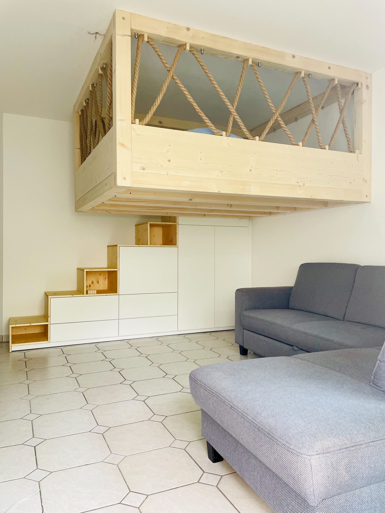 Hochbett aus Holz über einem Wohnzimmer mit Stufen nach oben. Weiße und graue Möbel.