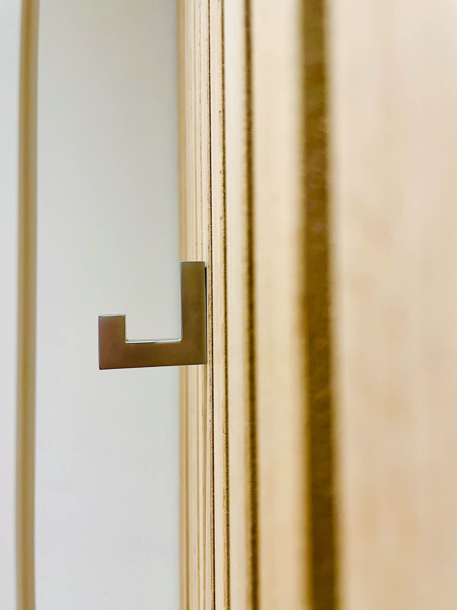 Nahaufnahme eines L-förmigen Metallhakens an einer Schranktür aus hellem Holz. Im Hintergrund ist eine weiße Wand zu sehen.