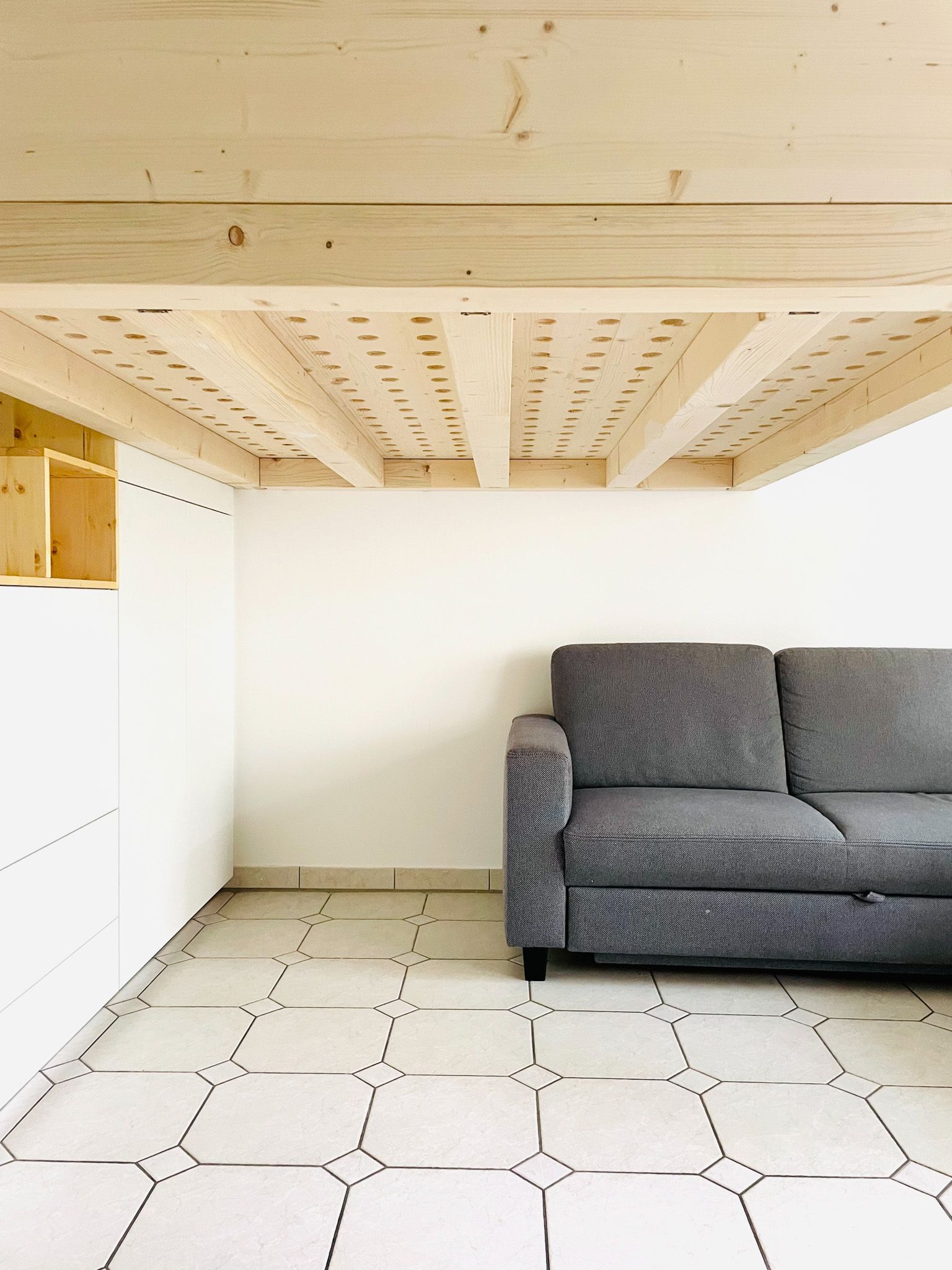 Graues Sofa vor einer weißen Wand und gemustertem Boden, mit hölzernen Deckenbalken darüber und einem Regal an der Seitenwand.