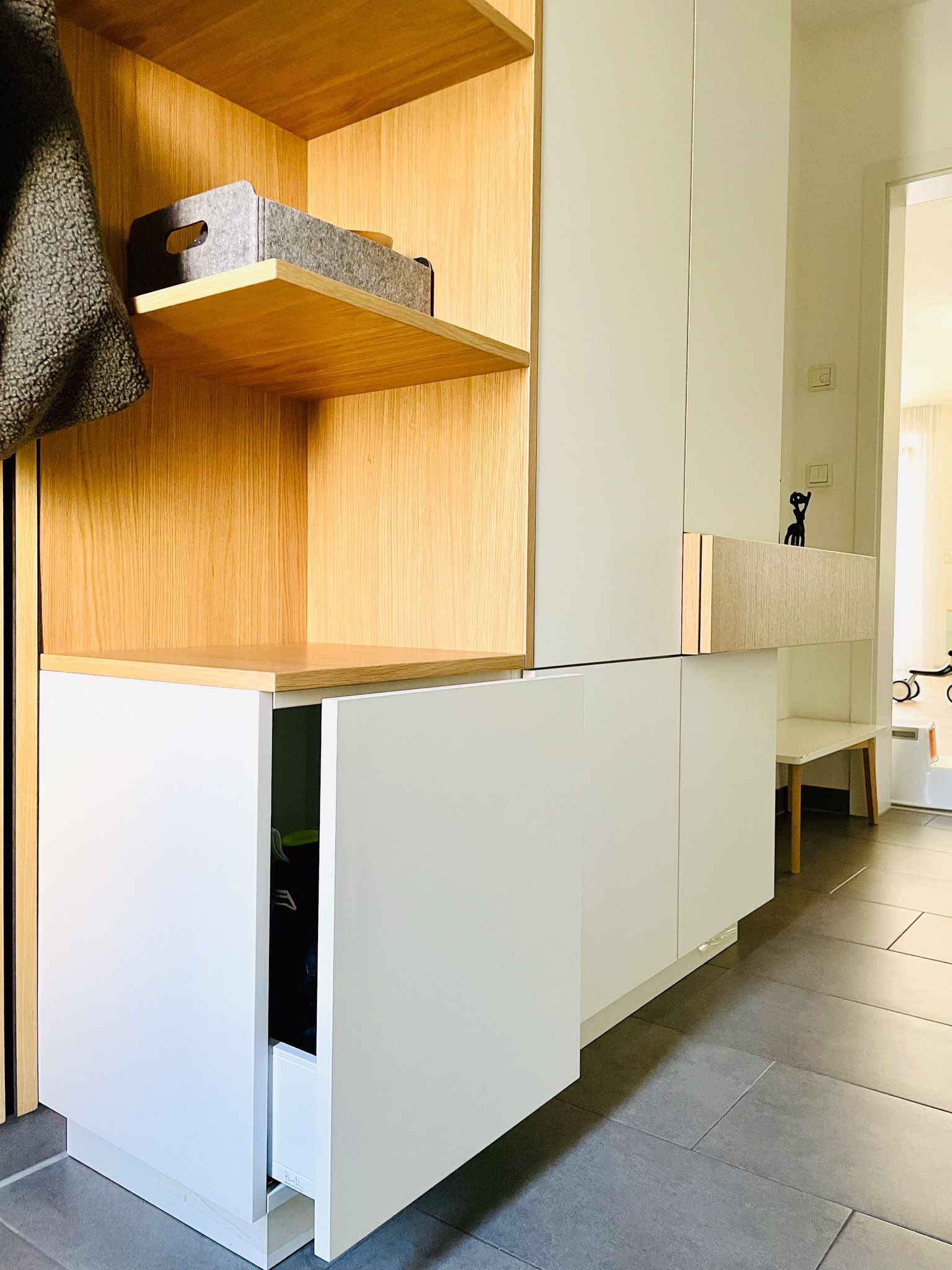 Ein moderner Eingangsbereich mit Stauraum in Weiß und Holz, Regalen und einer offenen Schublade. Grauer Bodenbelag.