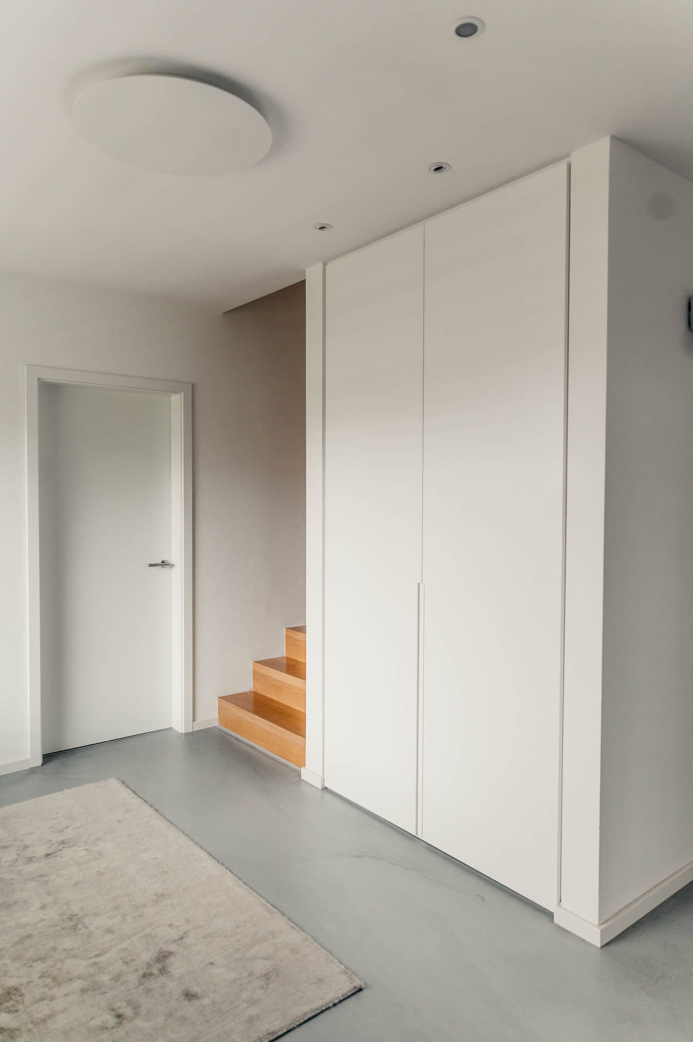 Strahlend weißer Flur mit Schrank, Tür und Holztreppe. Auf dem grauen Boden liegt ein Teppich, an der Decke ist eine runde Leuchte zu sehen.