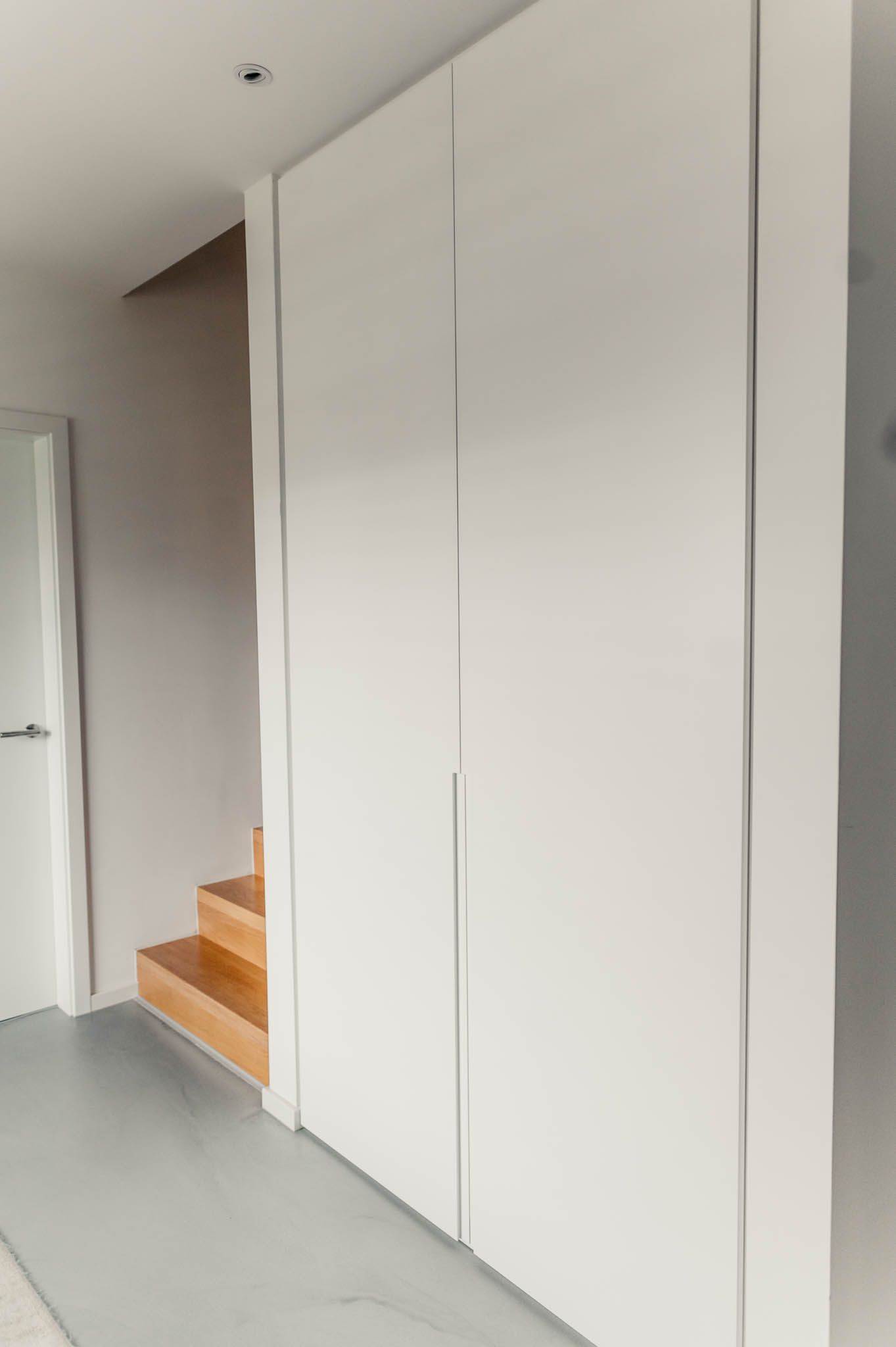 Weißer Einbauschrank mit vertikalen Linien, neben Holztreppen und einer weißen Tür, in einem hellen Flur.