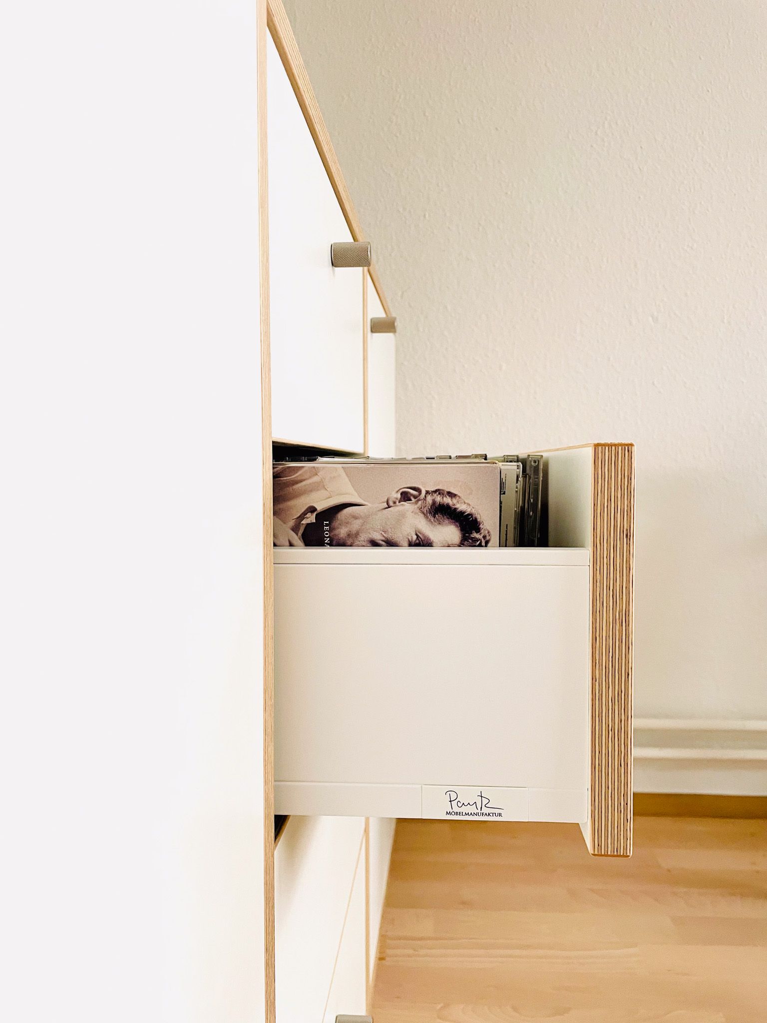 Weißer Schrank mit einer teilweise geöffneten Schublade, die den Inhalt freigibt. Die Schublade hat eine natürliche Holzkante.