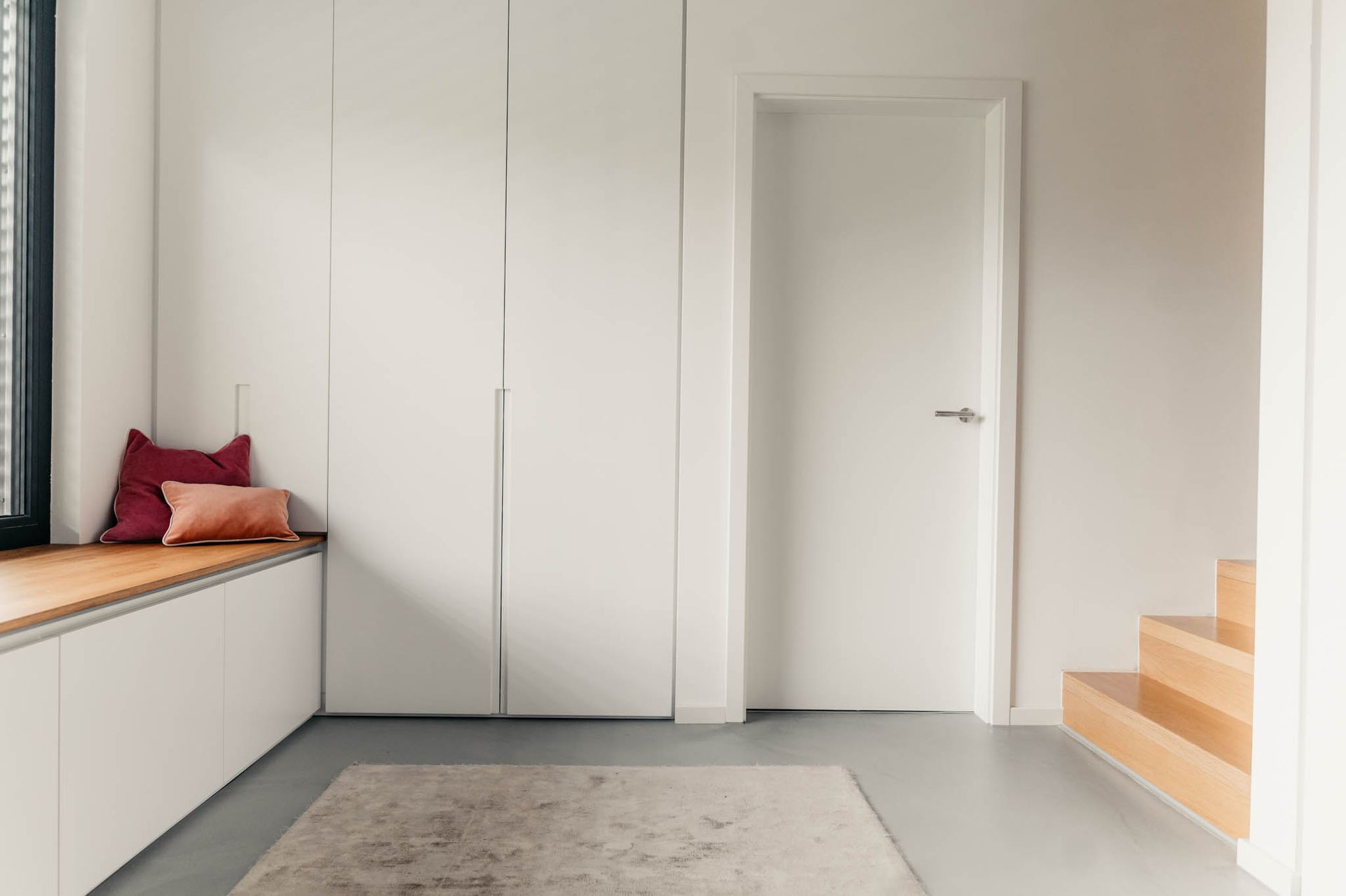Ein modernes Interieur mit einem weißen Kleiderschrank und einer weißen Tür, einem Fenstersitz mit Kissen und einem Teppich.