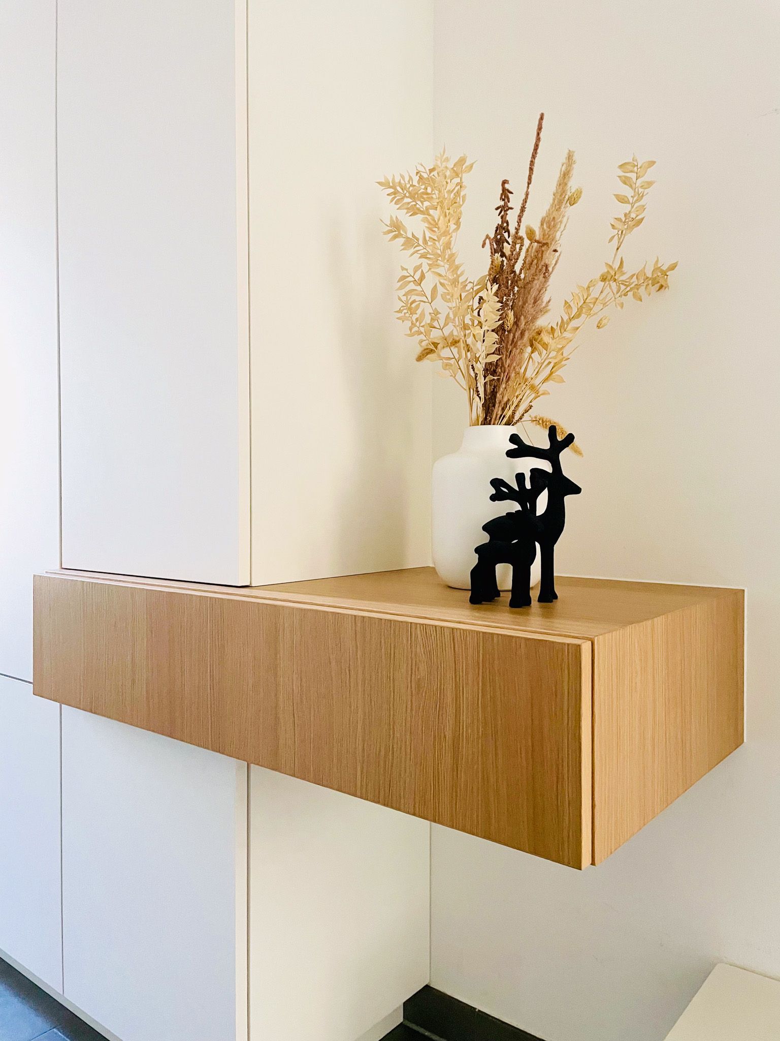 Schwebendes Holzregal mit weißer Vase mit getrockneten Blumen und schwarzer Hirschskulptur.